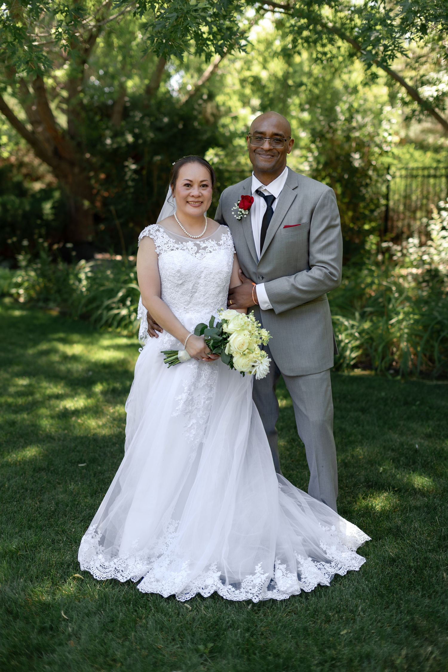 Interracial bride and groom pose together in lush garden, bride holding white bouquet in elegant lace wedding dress.
