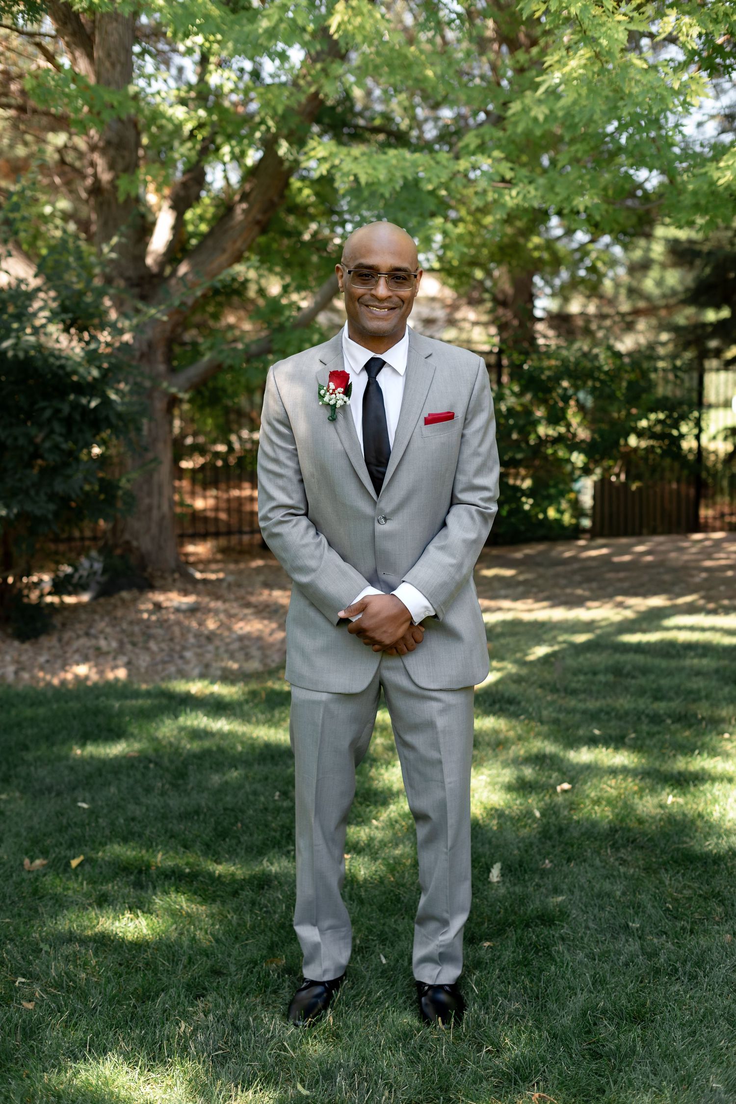 Groom in grey suit with red boutonniere and pocket square standing on green lawn.