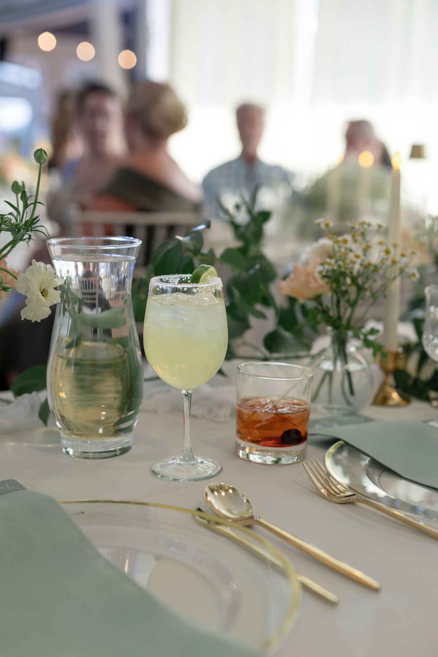 Elegant wedding reception table with gold cutlery, margarita cocktail, whiskey glass, and floral arrangements.