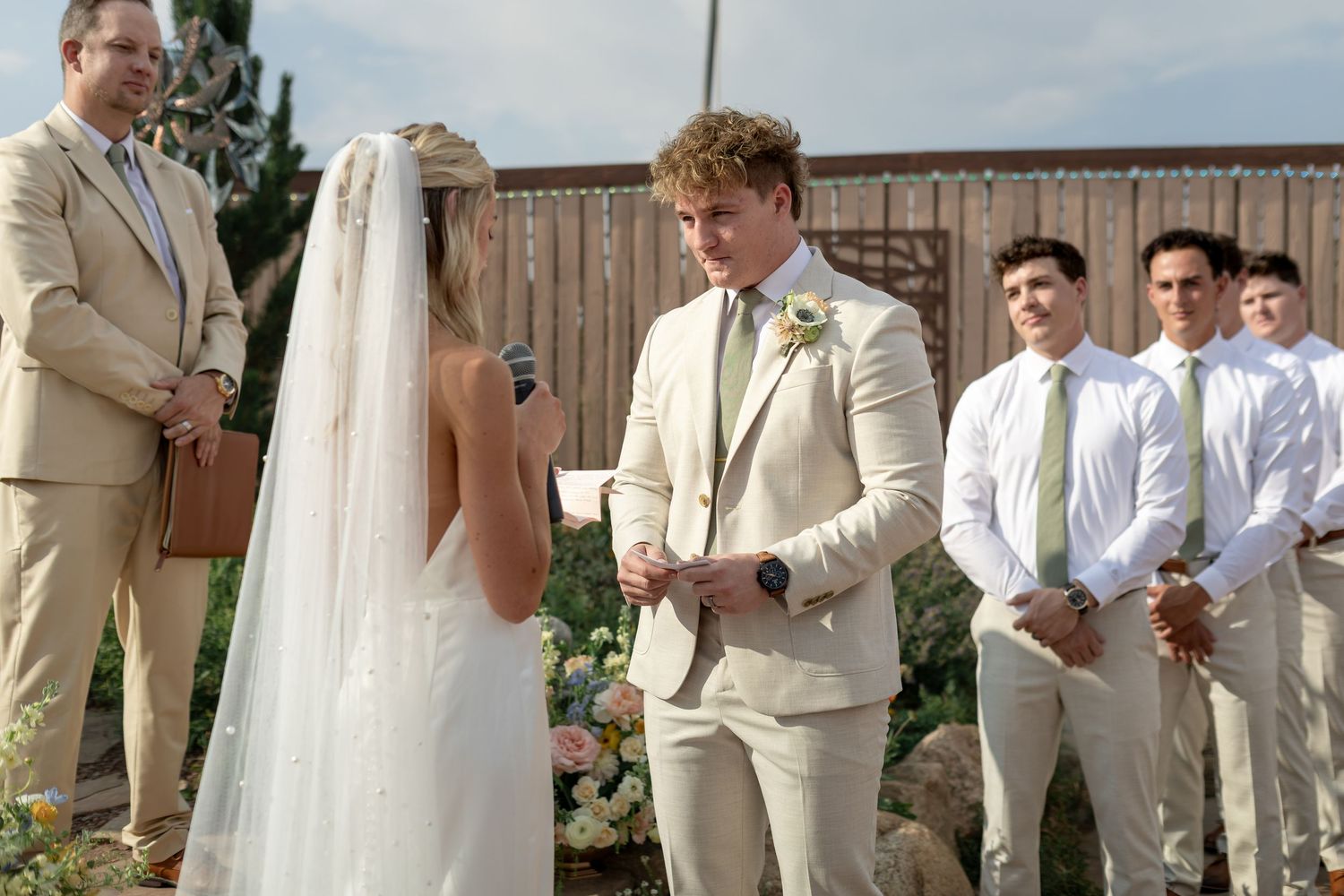Groom in tan suit reads vows to bride at outdoor wedding ceremony with groomsmen standing behind him.