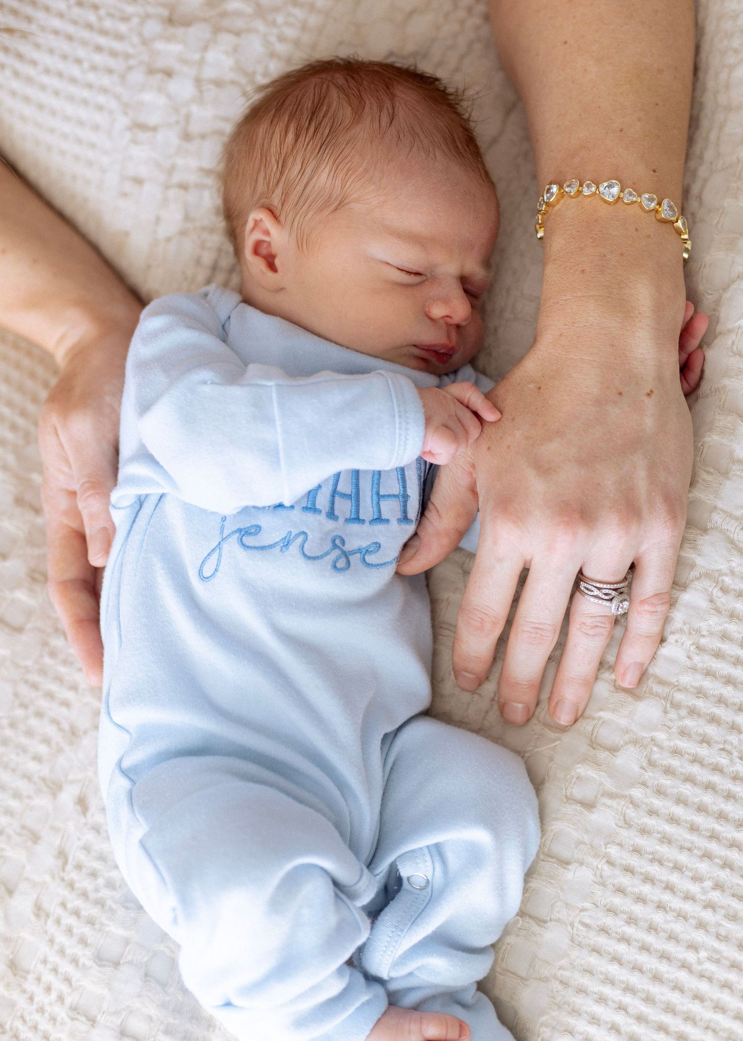 Sleeping infant in light blue outfit resting on white textured surface with caregiver's hands nearby.
