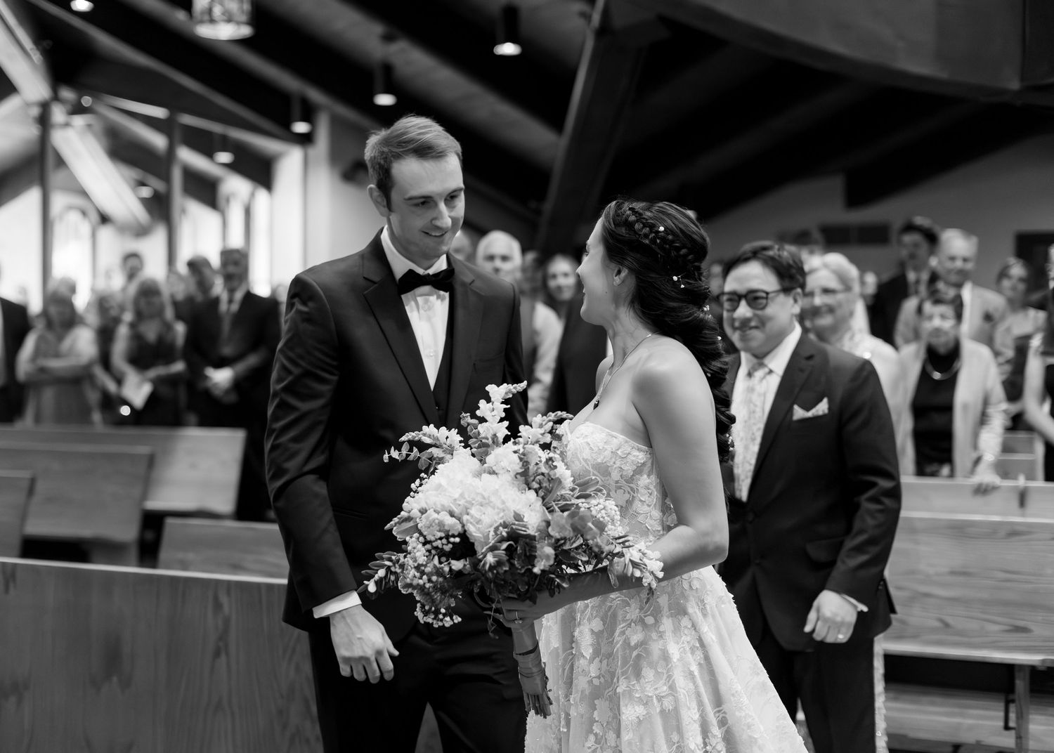 Black and white wedding photo of bride holding bouquet and groom sharing a moment at the altar in a church.