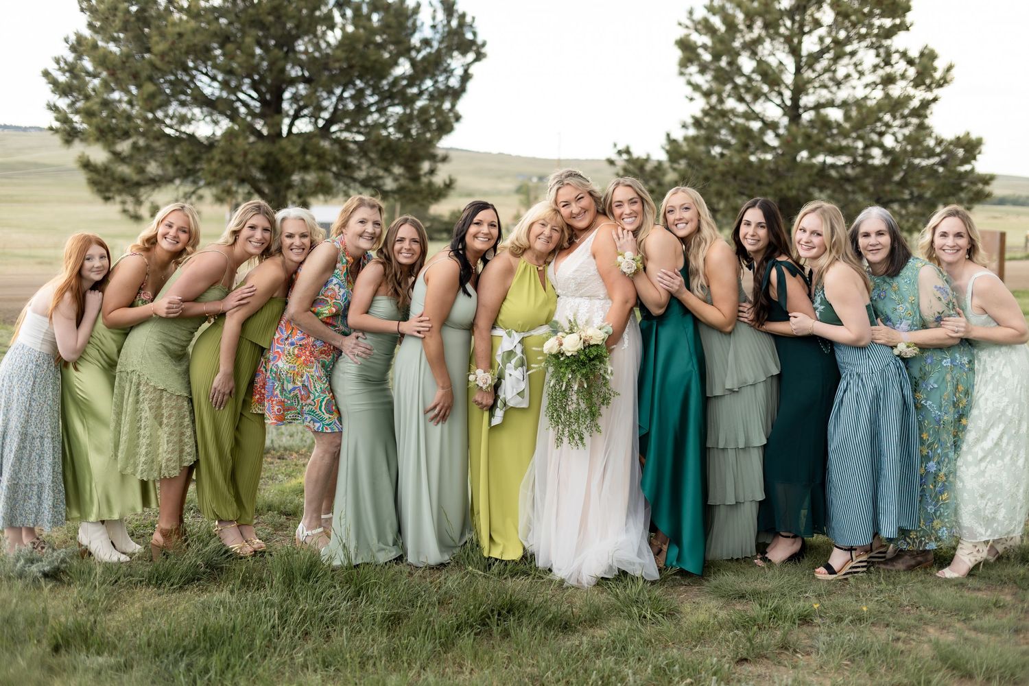 Bride in white gown poses with bridesmaids in mismatched green dresses outdoors on a scenic grassy hillside.