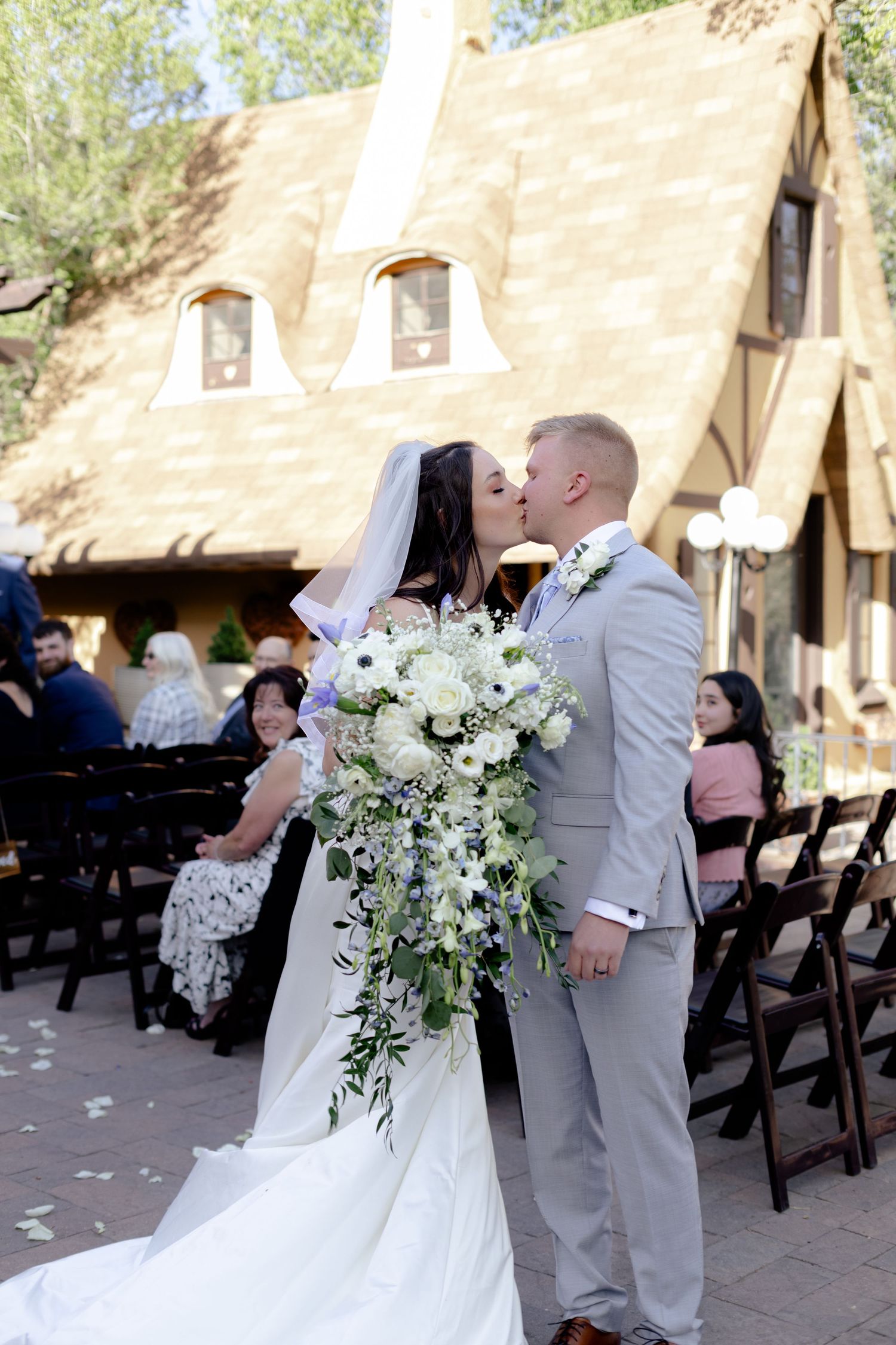 Bride and groom share first kiss at outdoor wedding ceremony beside a charming storybook chapel with guests seated behind.