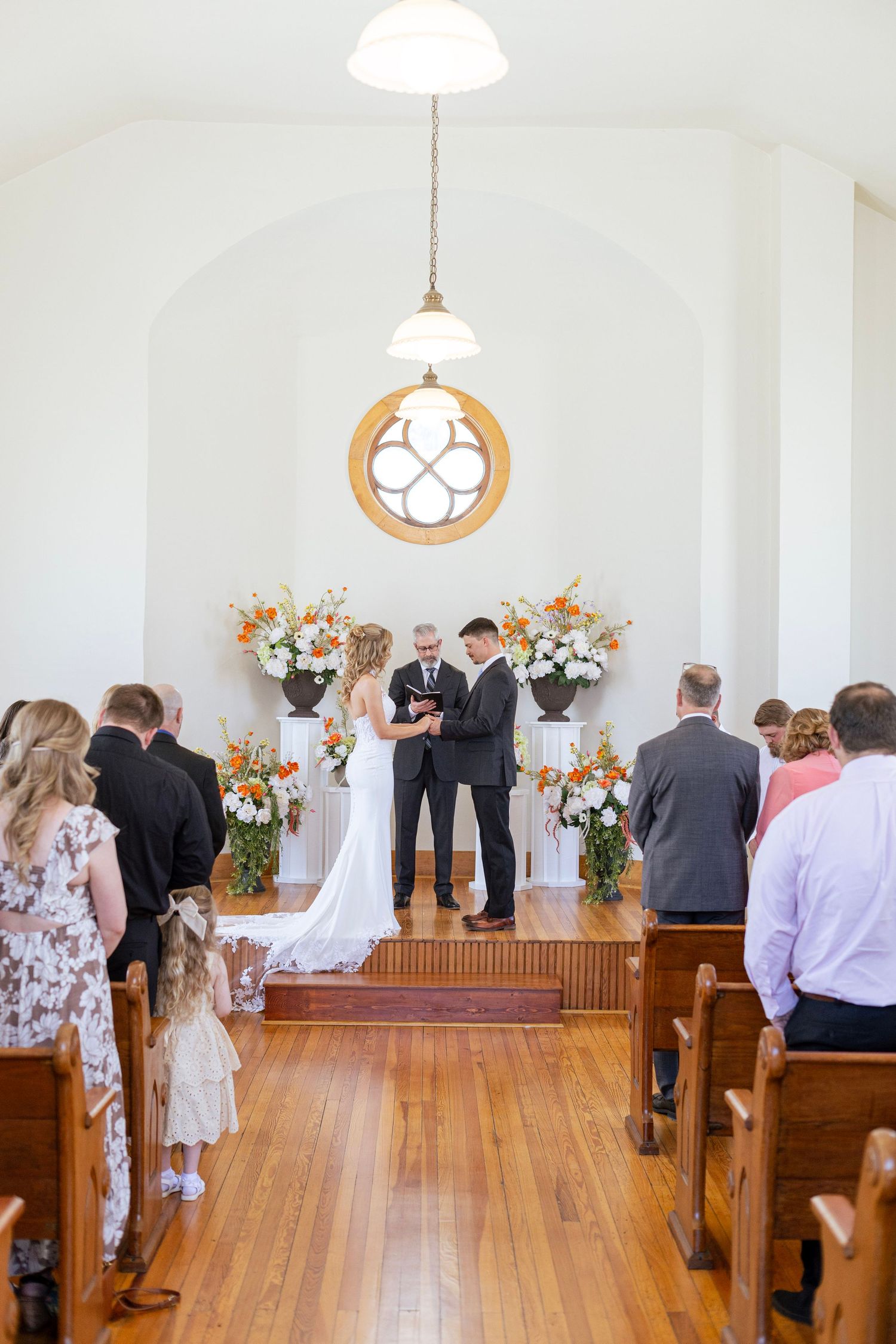 Bride and groom exchanging vows at chapel altar with orange floral arrangements and wooden pews.