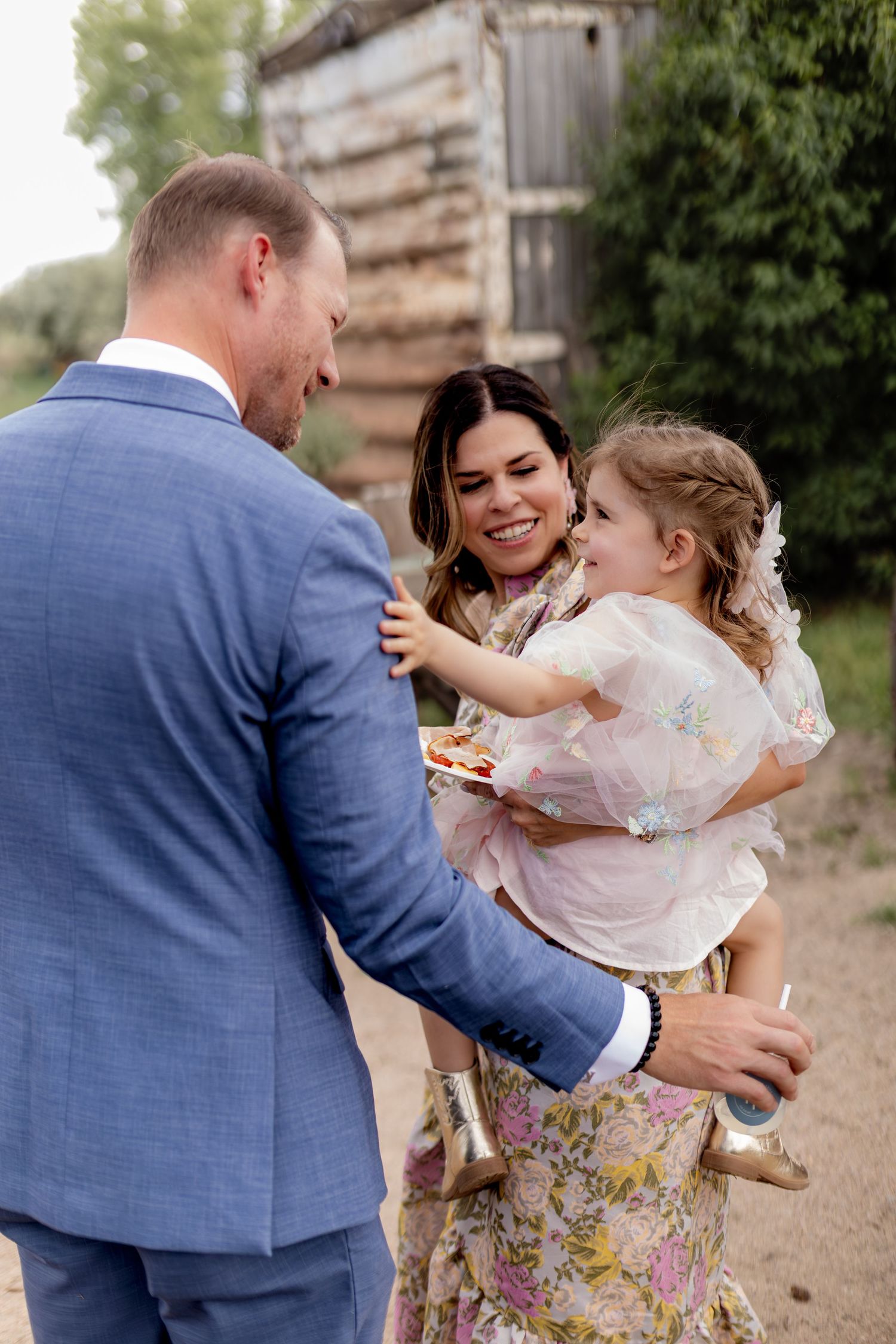 Couple in wedding attire smiling while holding young girl in floral dress at outdoor celebration.