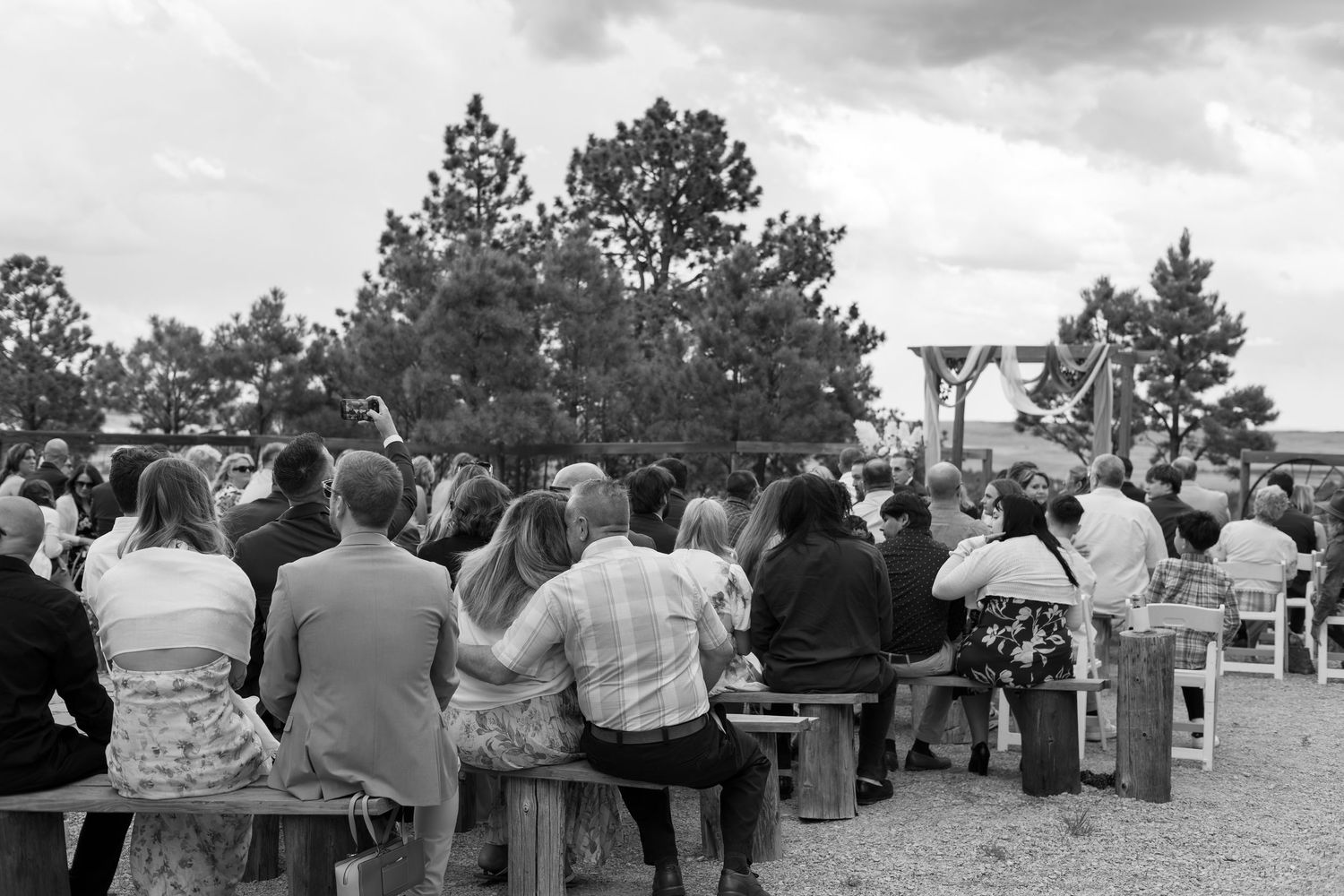 Guests seated on wooden benches at an outdoor wedding ceremony surrounded by trees in black and white.