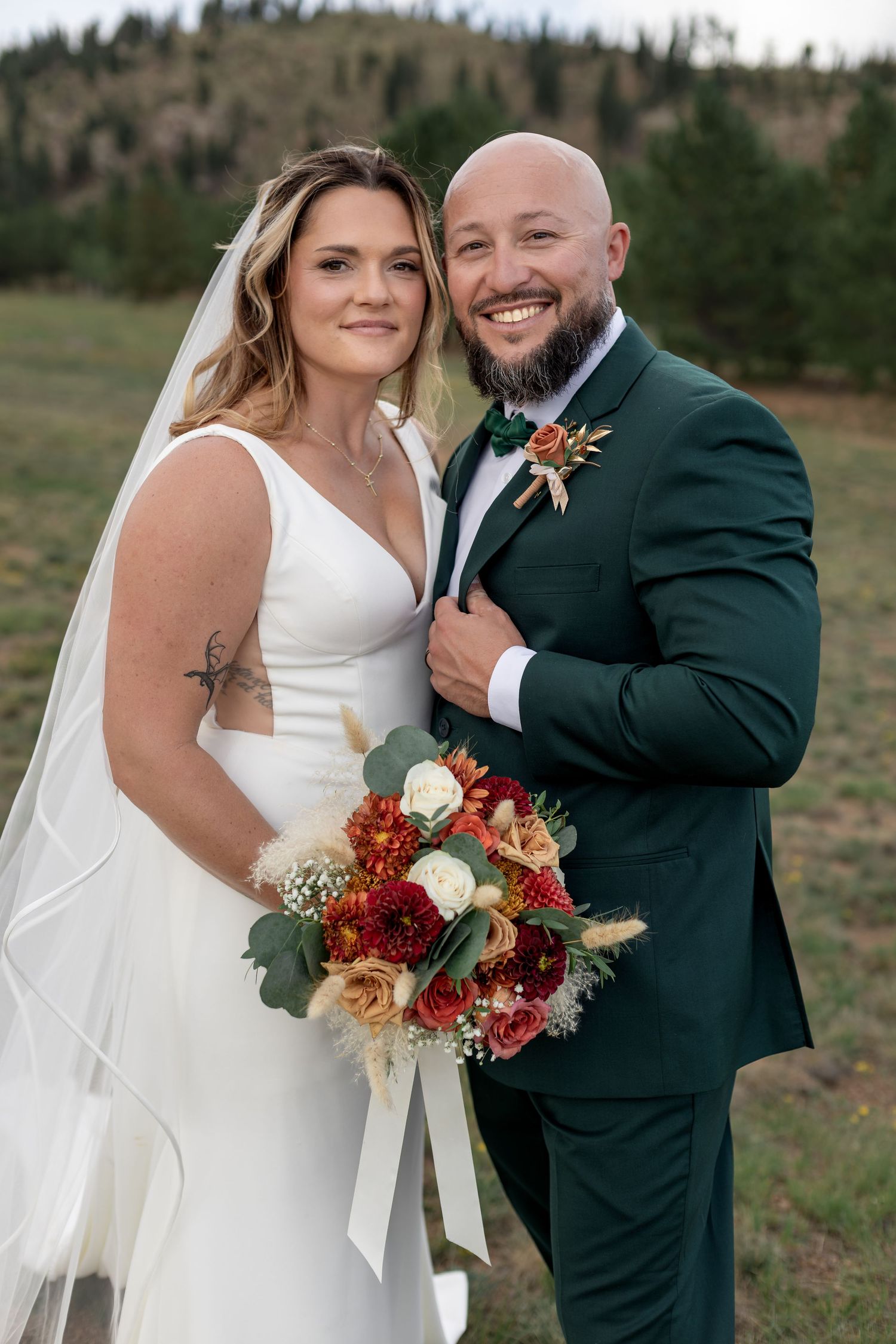 Bride in white gown holding red bouquet poses with bald groom in green suit at outdoor mountain wedding.