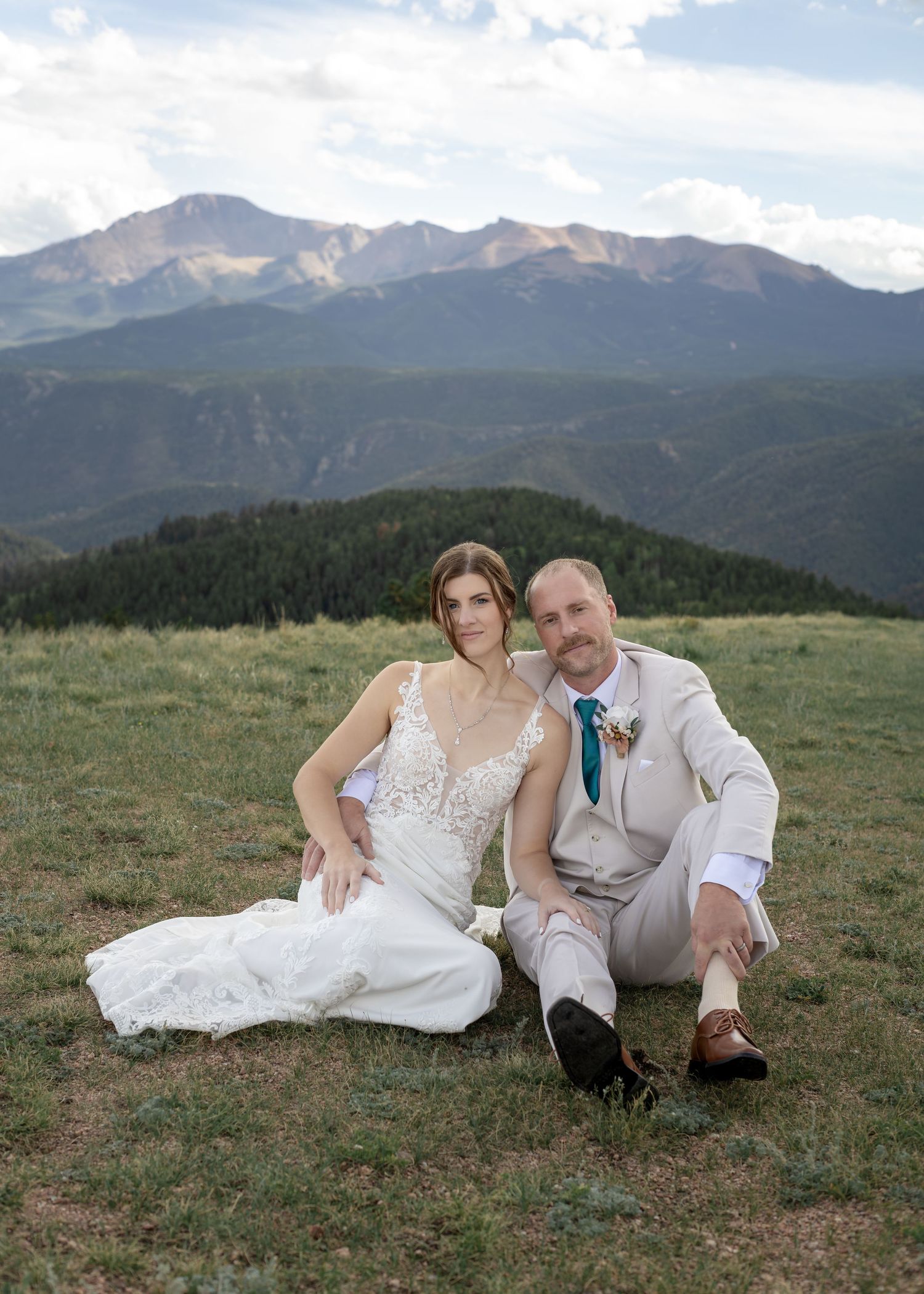 Bride and groom sitting on mountain meadow with Rocky Mountain peaks backdrop in Colorado wedding portrait.