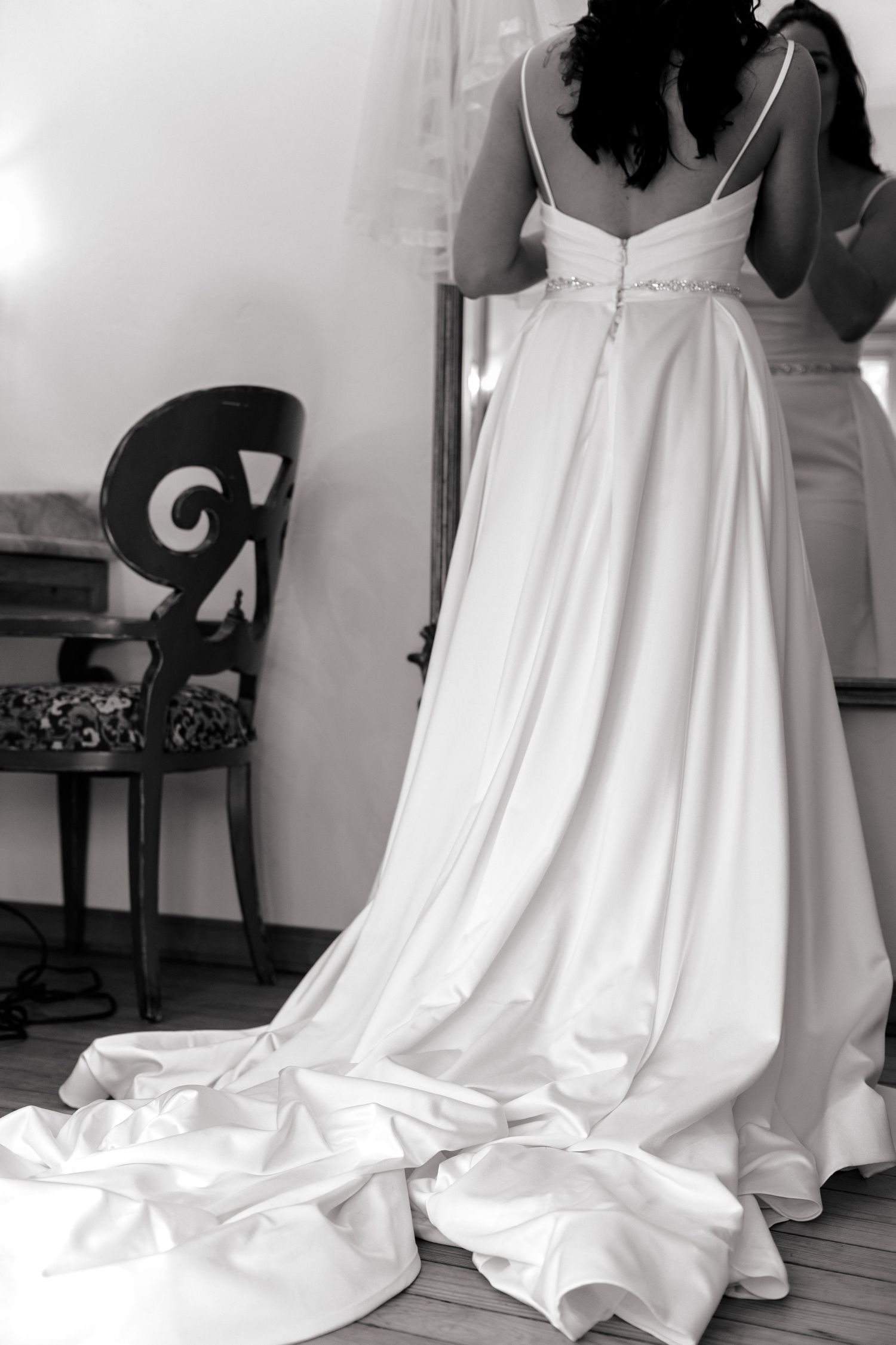 Black and white photo of a bride in an elegant satin wedding gown with a long train, viewed from behind.