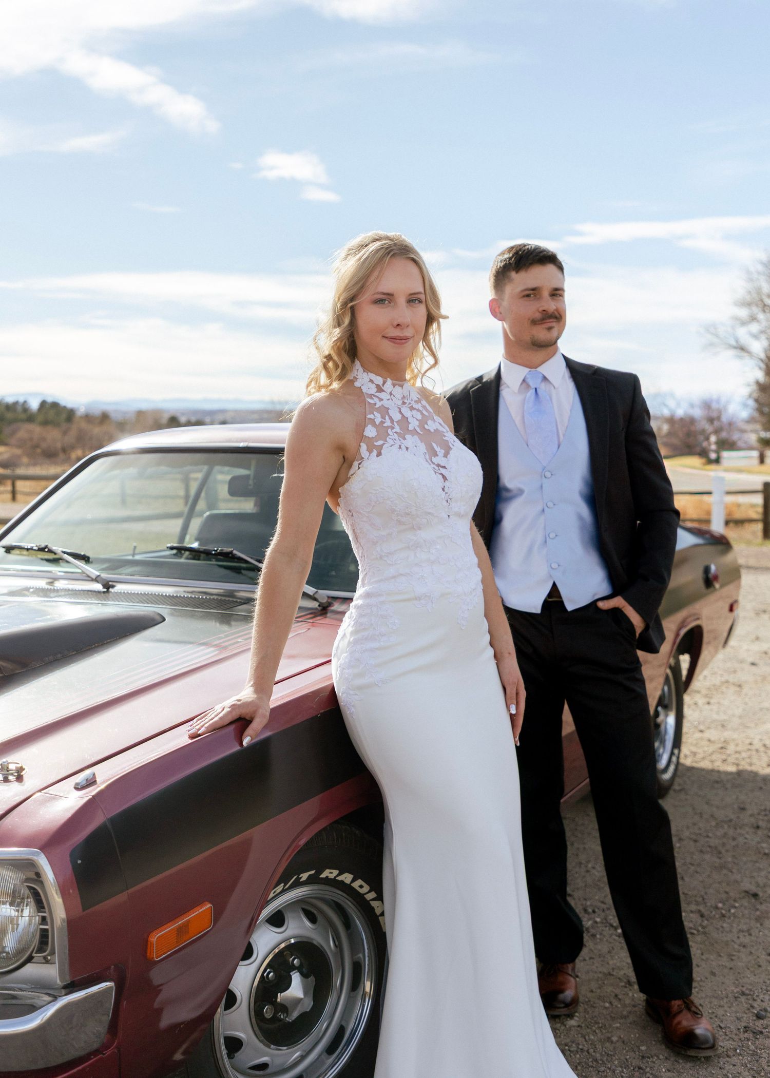 Bride in halter lace gown and groom posing beside vintage muscle car under blue sky.