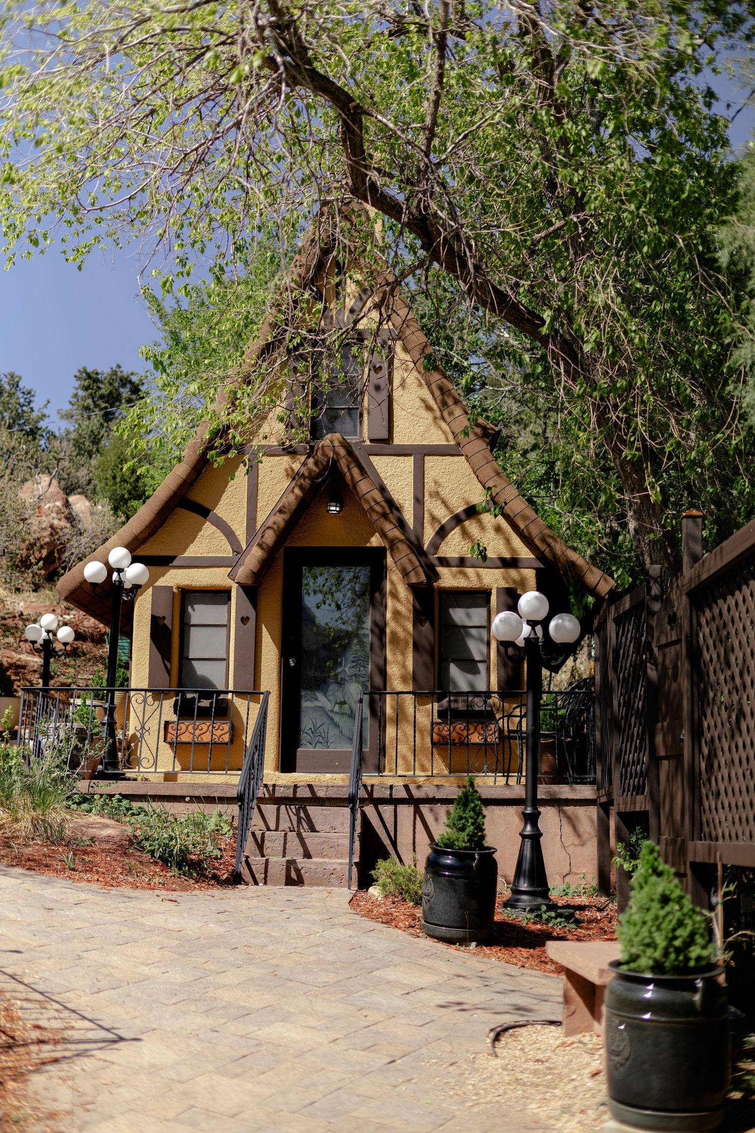 Charming Tudor-style cottage with steep gabled roof, globe lights, and lush trees in a garden setting.