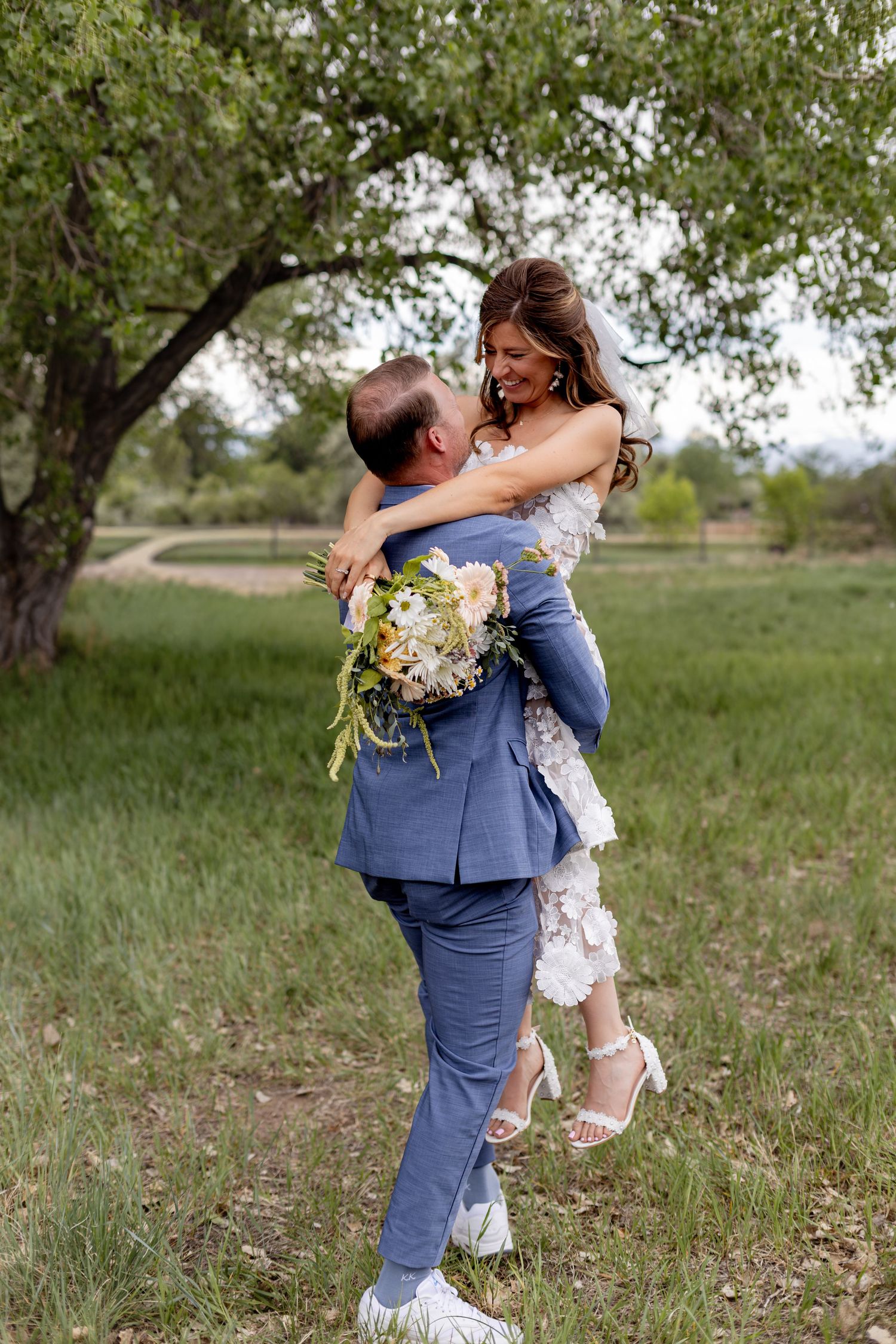 Groom lifting smiling bride in floral lace dress outdoors in a green meadow beneath a large tree.