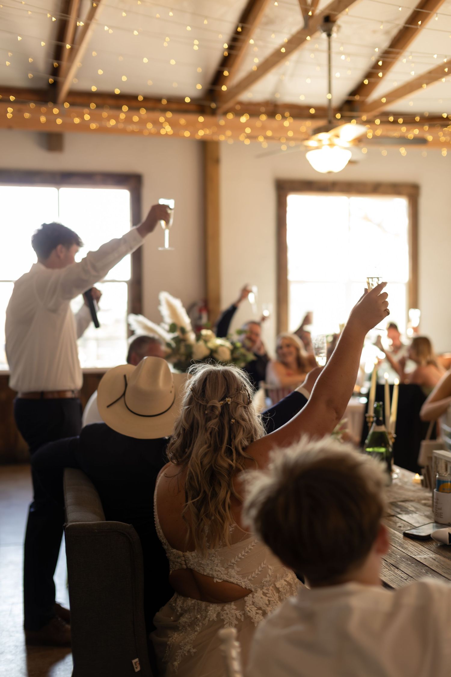 Wedding reception toast with guests raising glasses under string lights in rustic barn venue.
