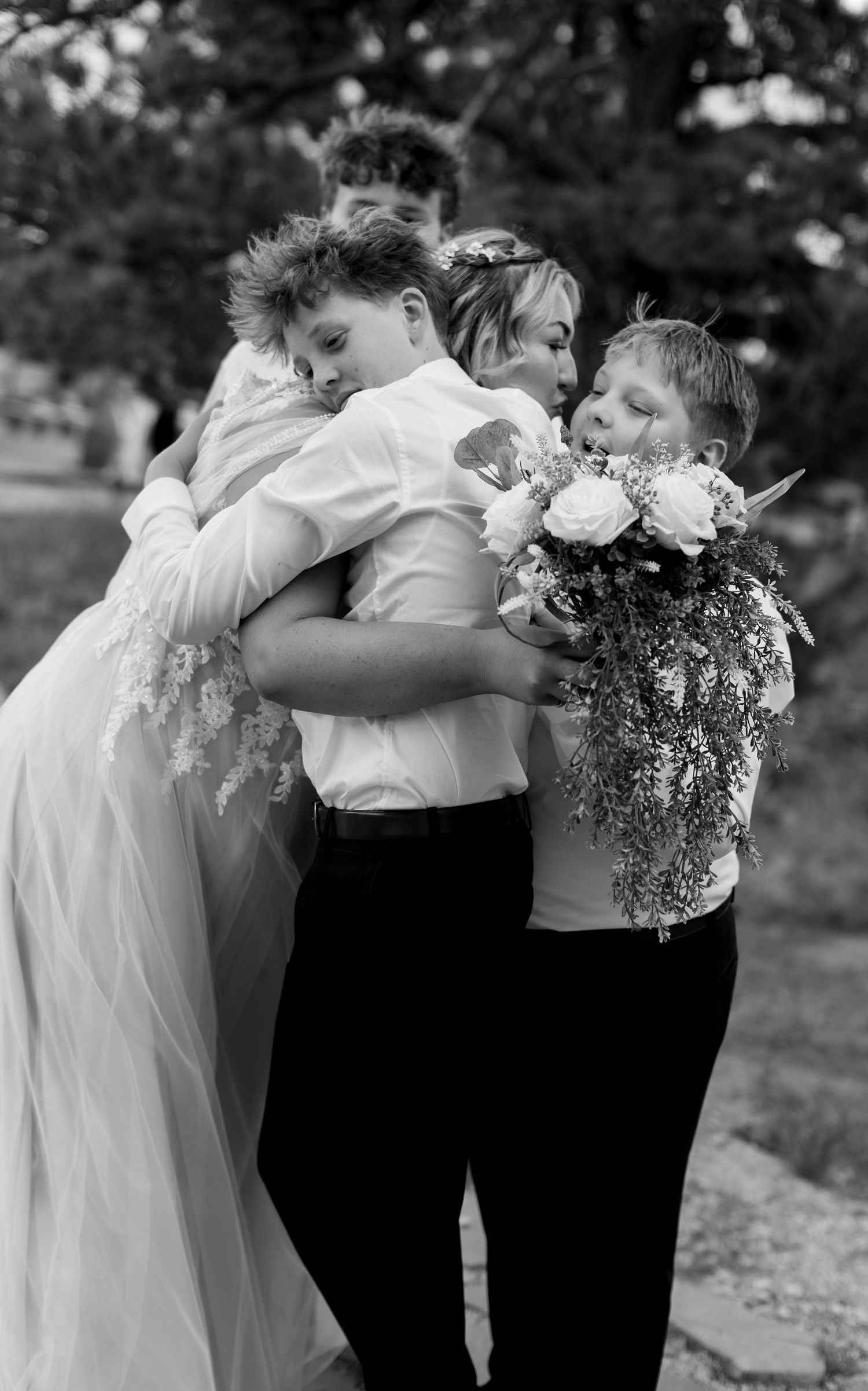 Bride in white gown embraced by children holding floral bouquet in black and white photo.