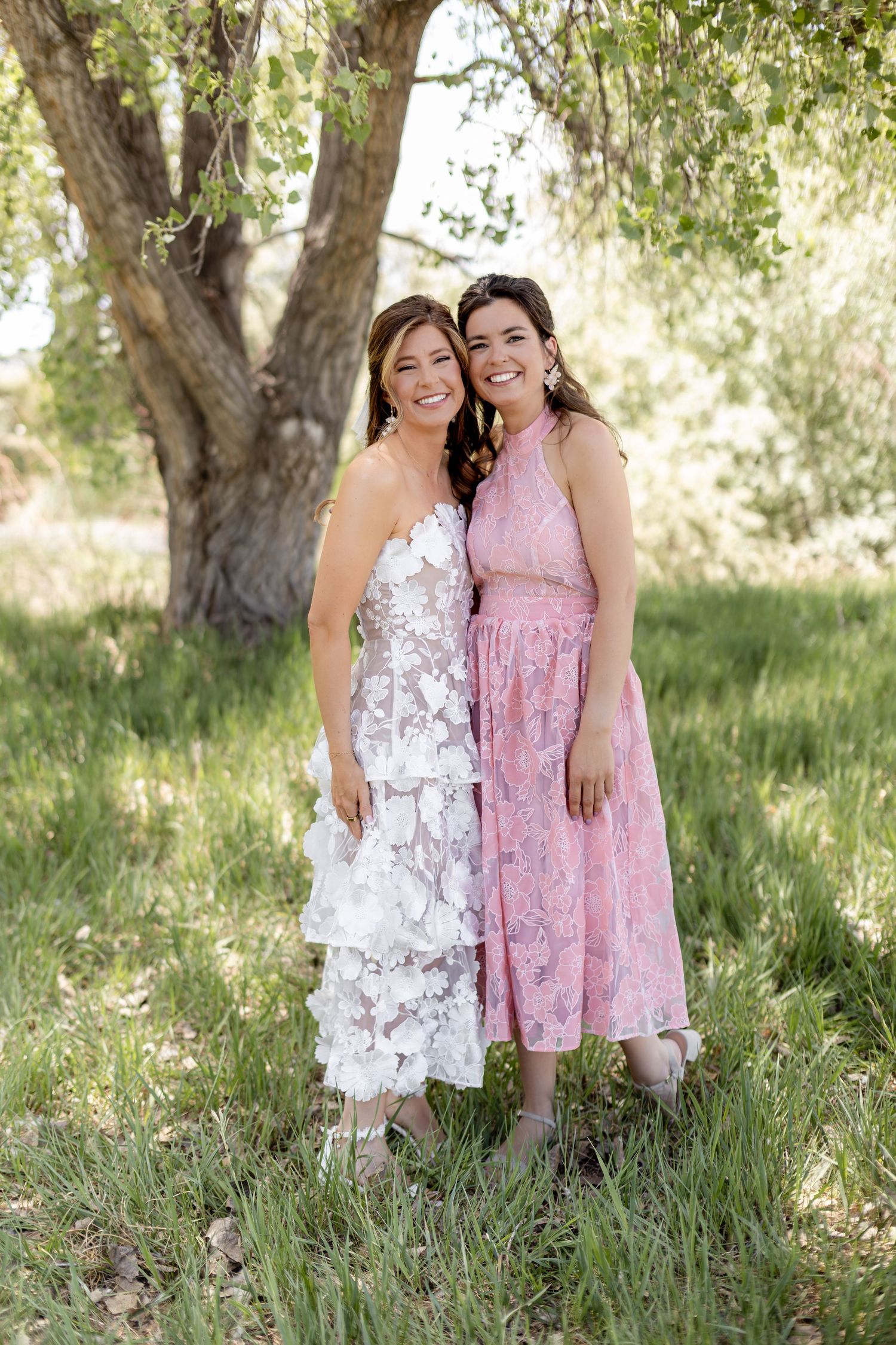Two smiling women posing together outdoors, one in a white floral bridal gown and one in a pink lace dress.