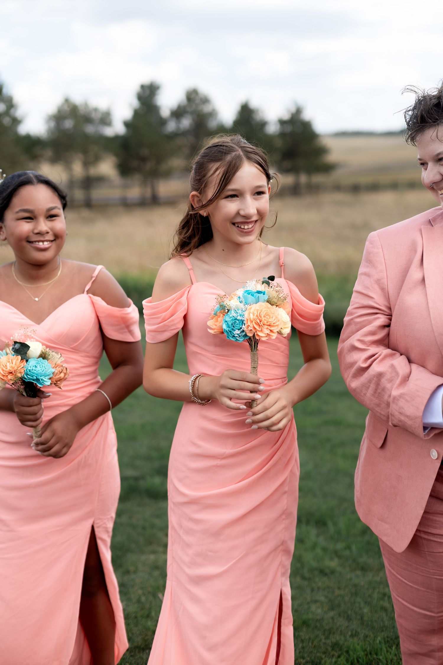Three bridesmaids in coral pink dresses walk outdoors, two holding colorful bouquets at a wedding.