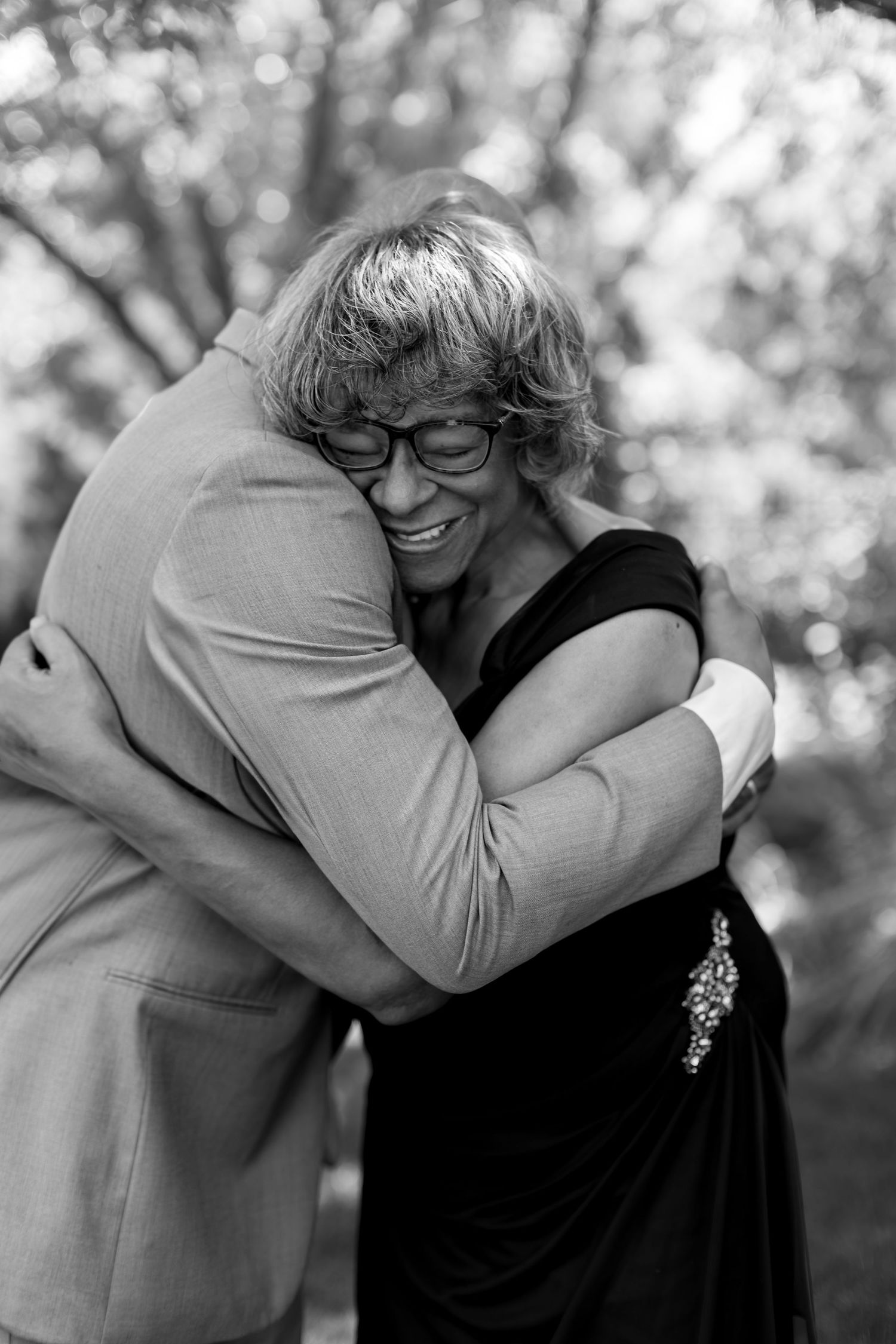 Black and white photo of two people sharing a warm embrace outdoors, woman in black dress smiling joyfully.
