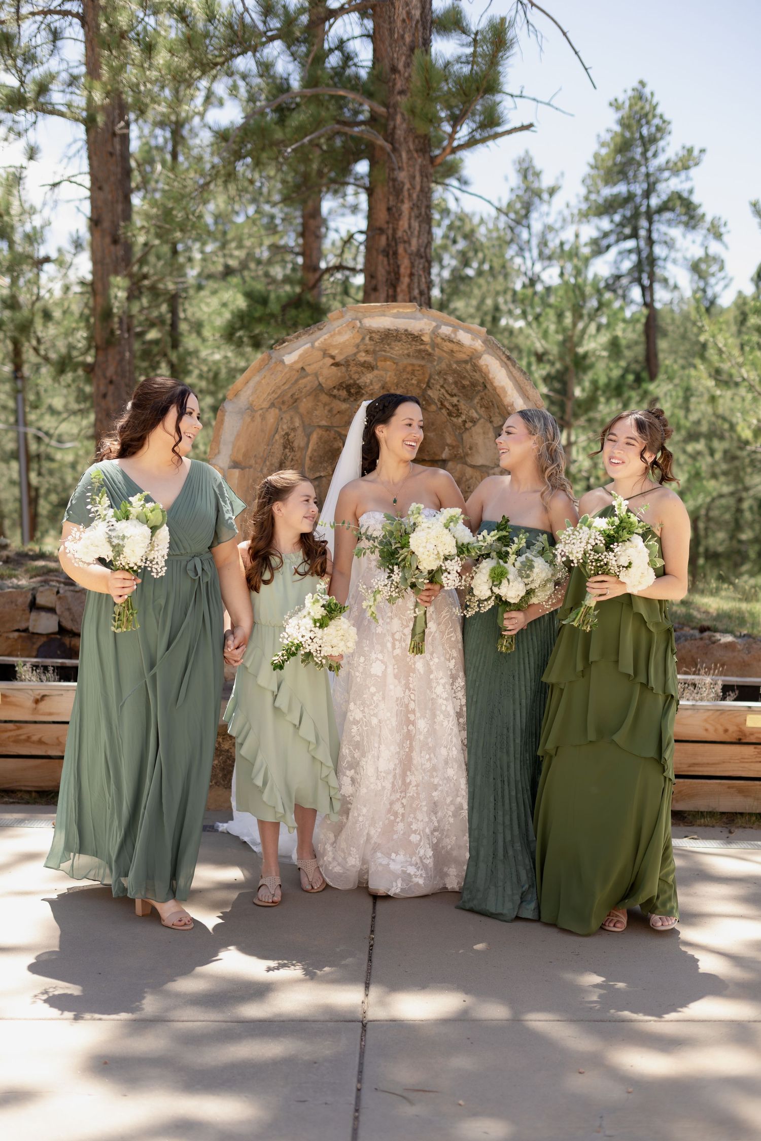 Bride in white gown with bridesmaids in green dresses holding bouquets outdoors at a forest wedding venue.
