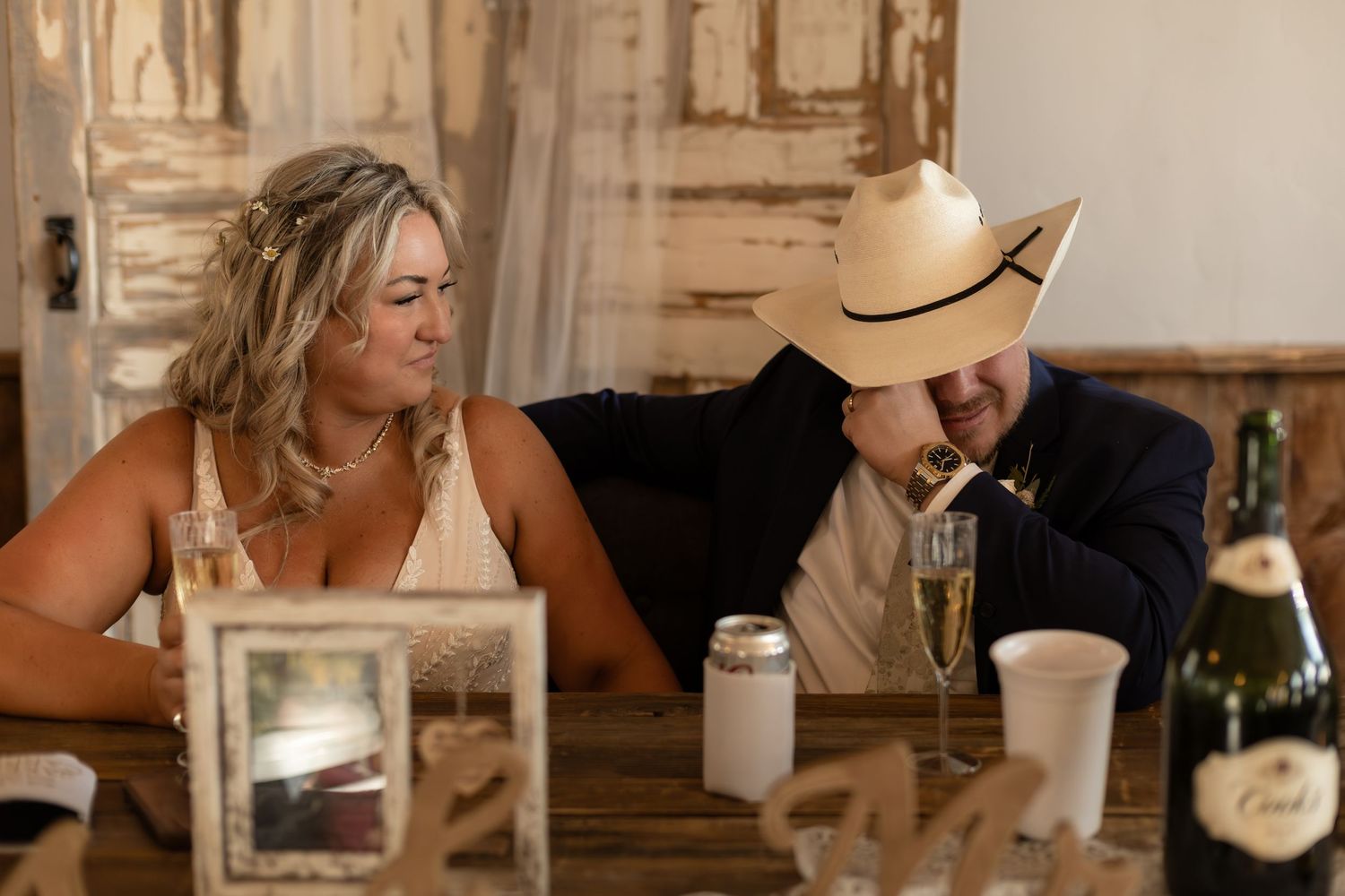 Bride and groom in cowboy hat sitting at rustic wooden table during wedding reception with champagne.