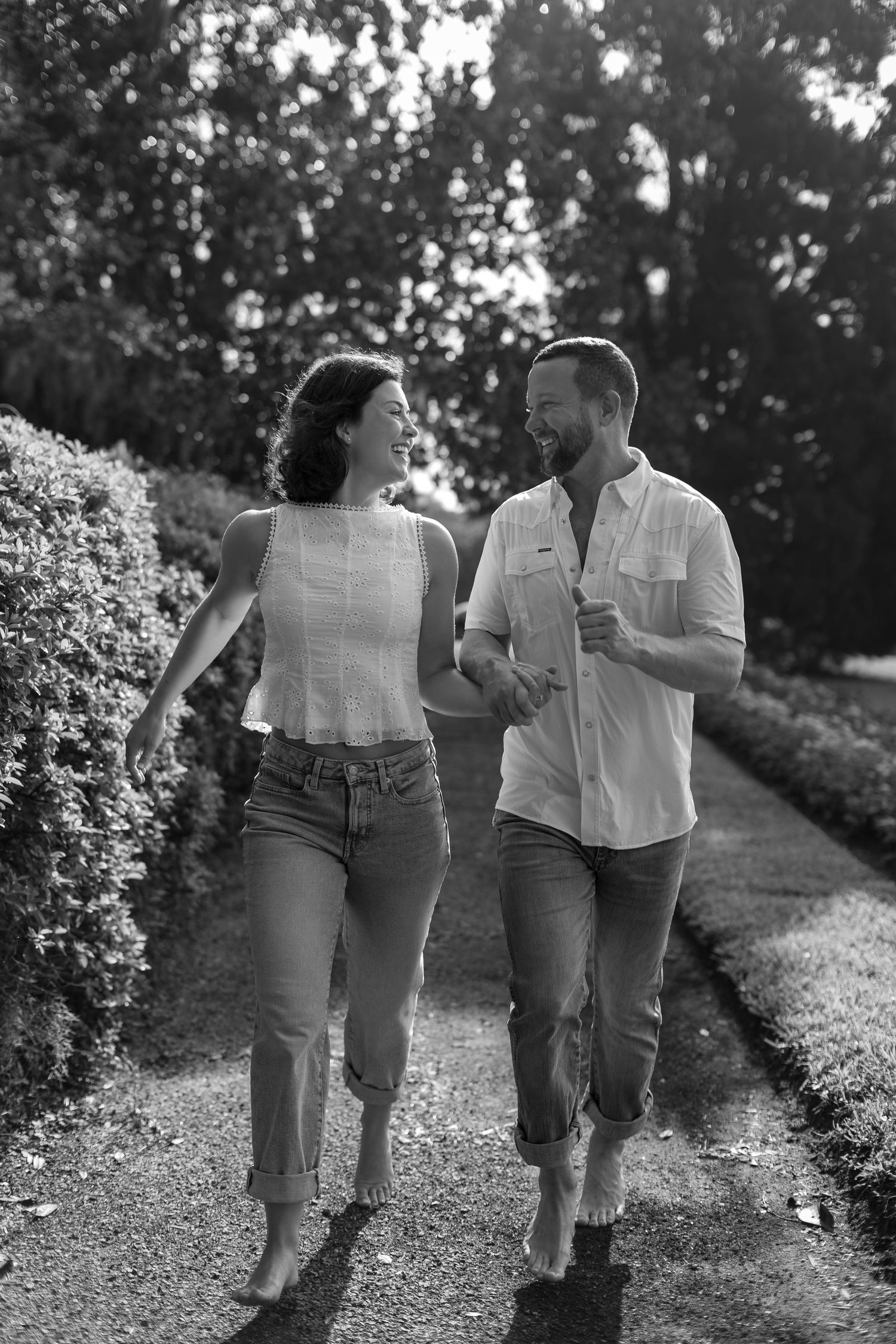 Black and white photo of a smiling couple walking hand in hand along a garden path surrounded by lush hedges and trees.
