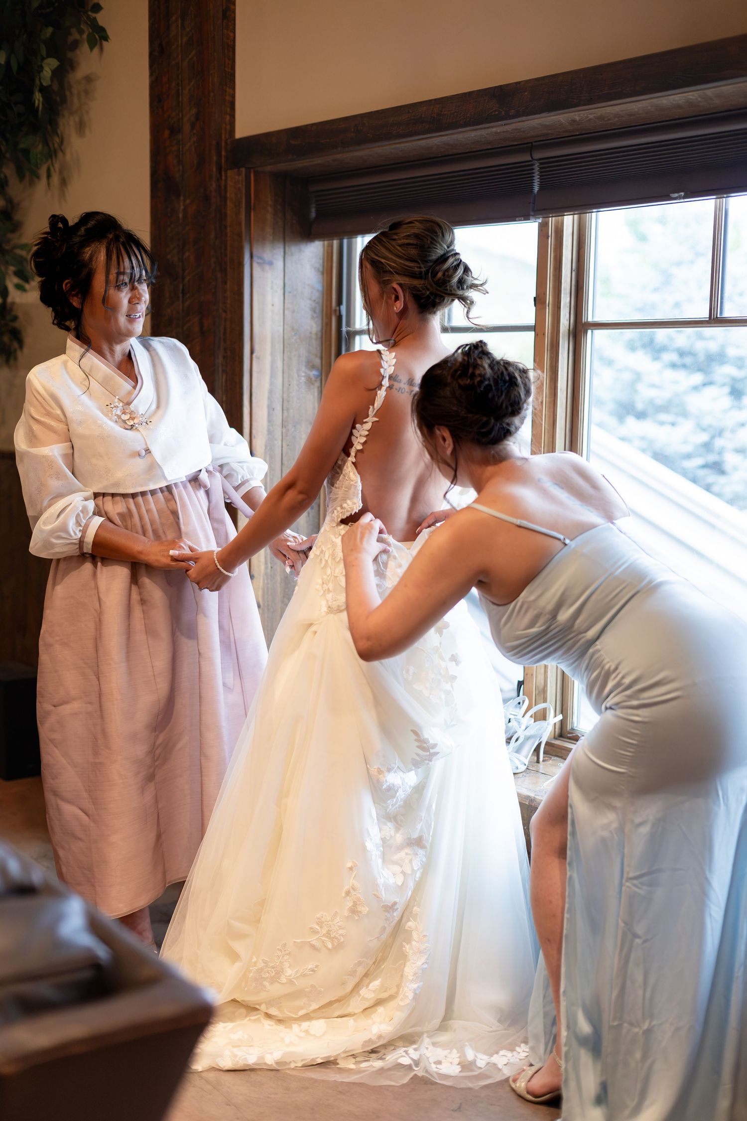 Bride being helped into white wedding gown by bridesmaid and mother near rustic wooden window.