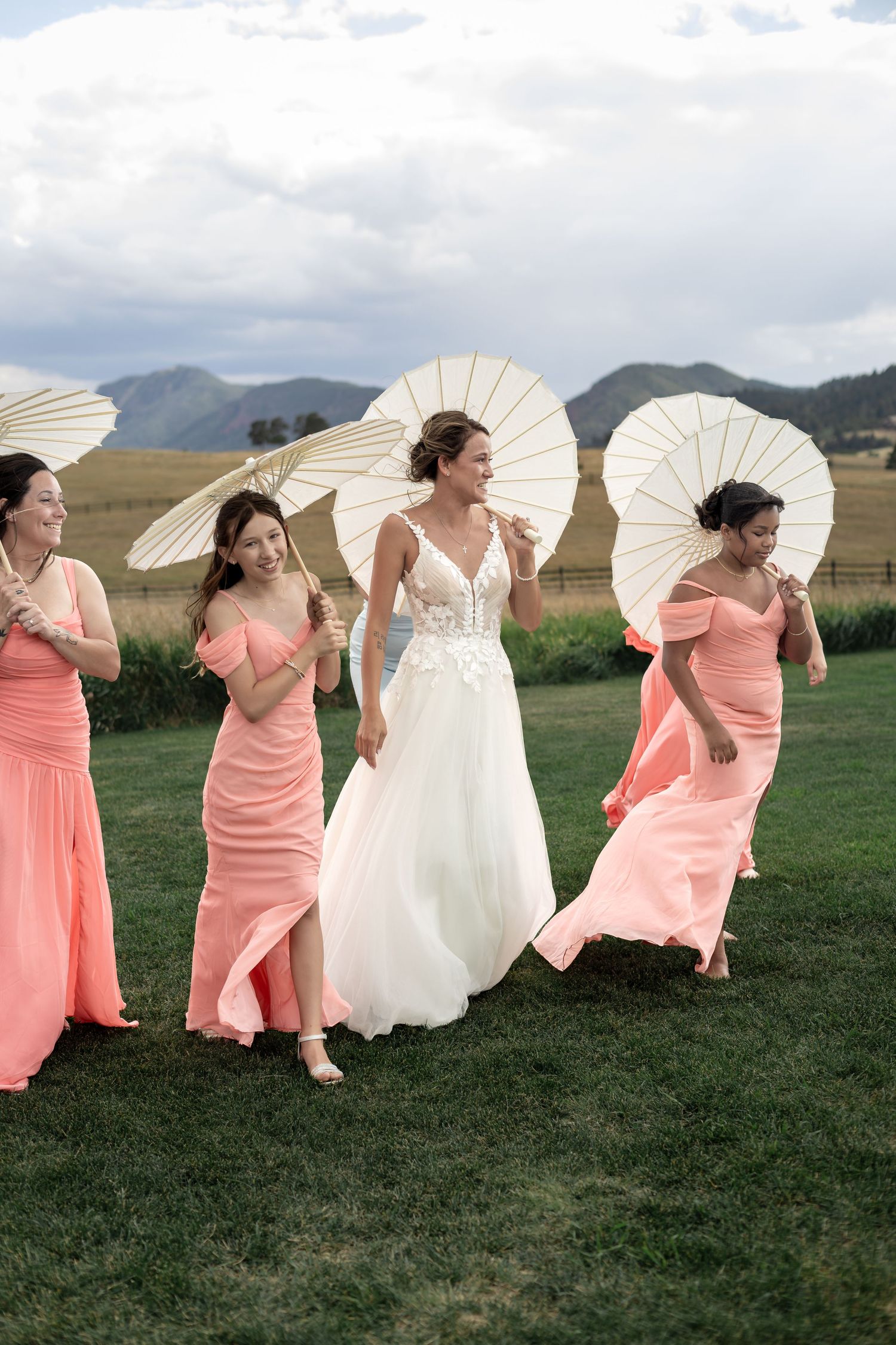 Bride and bridesmaids in coral dresses laughing and walking outdoors with white parasols.