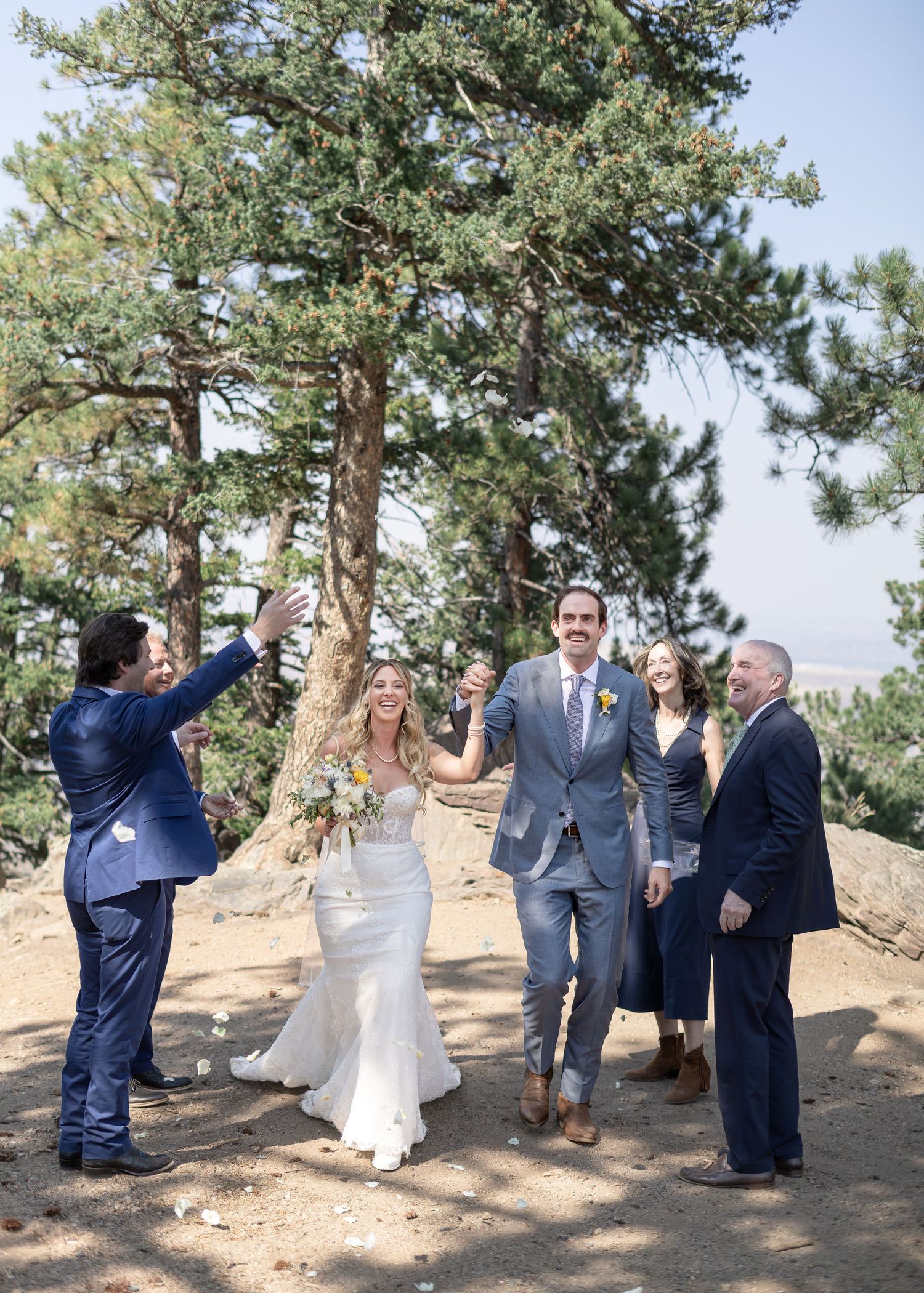 Bride and groom walk joyfully outdoors at mountain wedding ceremony surrounded by pine trees and smiling guests.