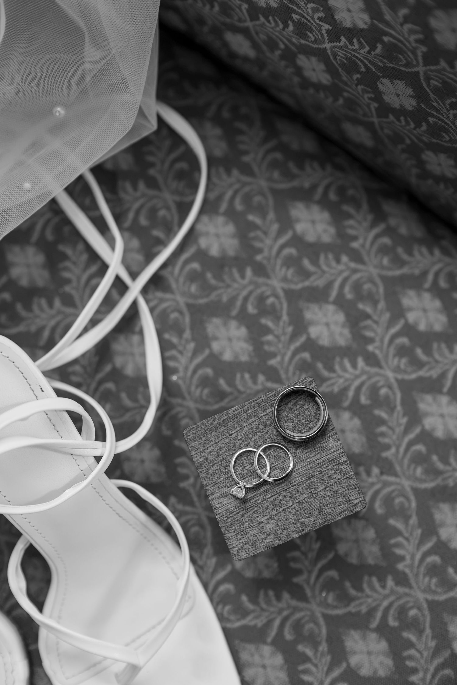 Black and white flat lay of wedding rings and bridal shoes on a patterned surface with veil.