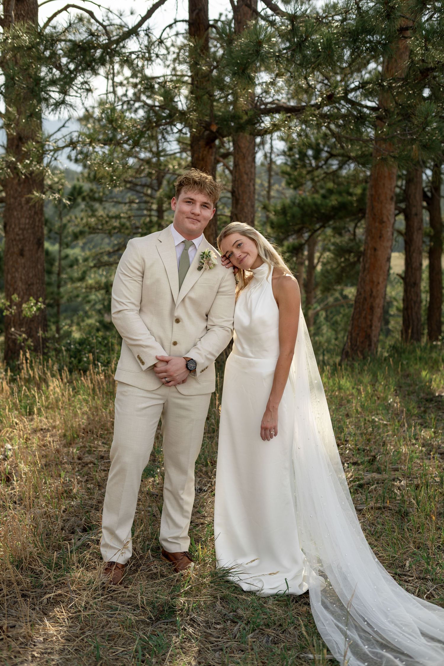 Bride and groom posing together in a pine forest, bride in white halter gown with veil, groom in cream suit.