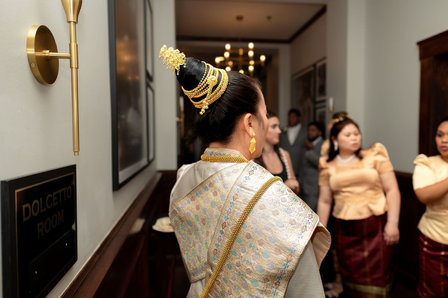 Woman in traditional Thai ceremonial dress with gold headdress stands in elegant hallway at formal event.