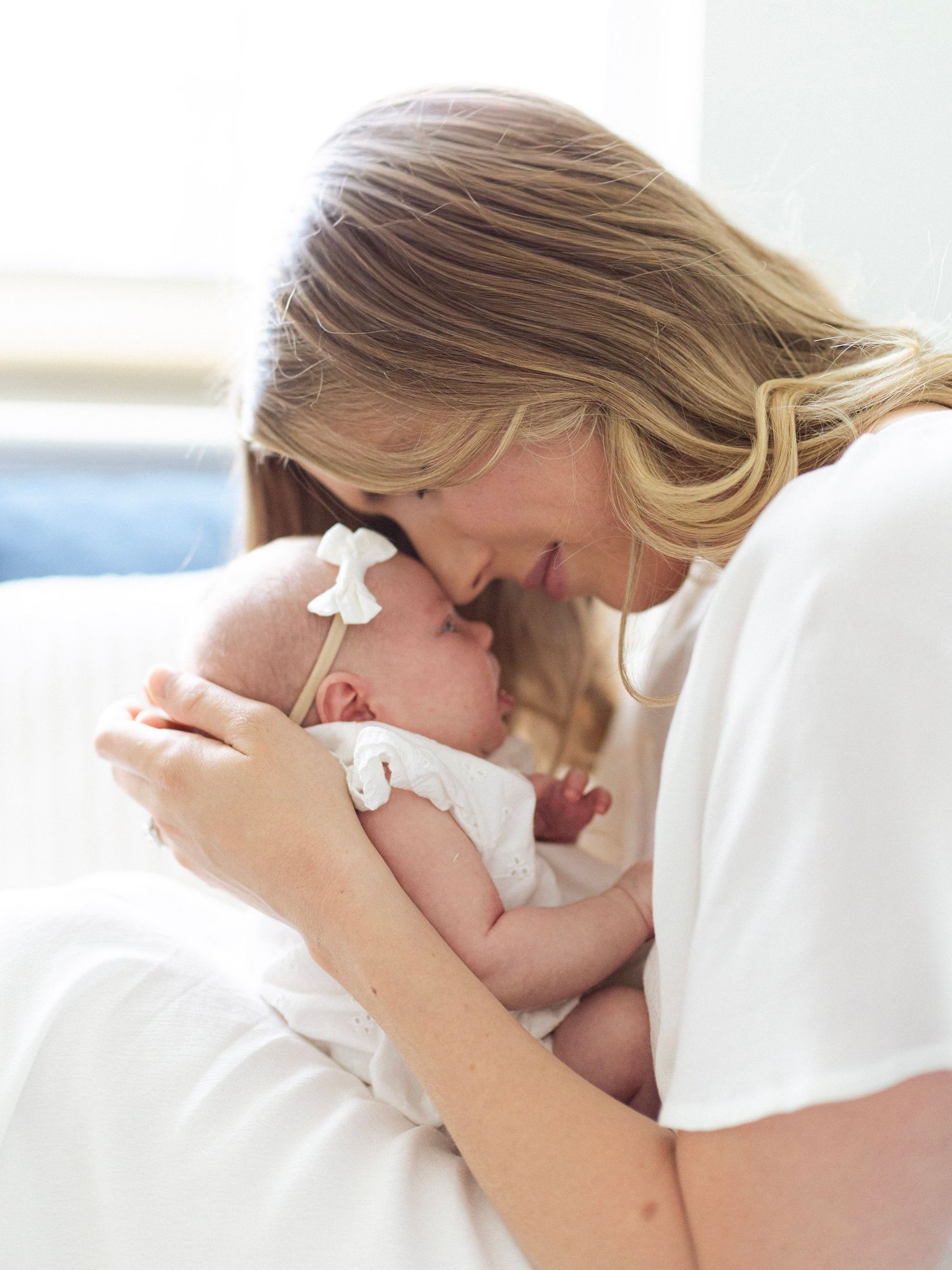 A blonde mother in white gently touches foreheads with her newborn baby dressed in white with a bow headband.