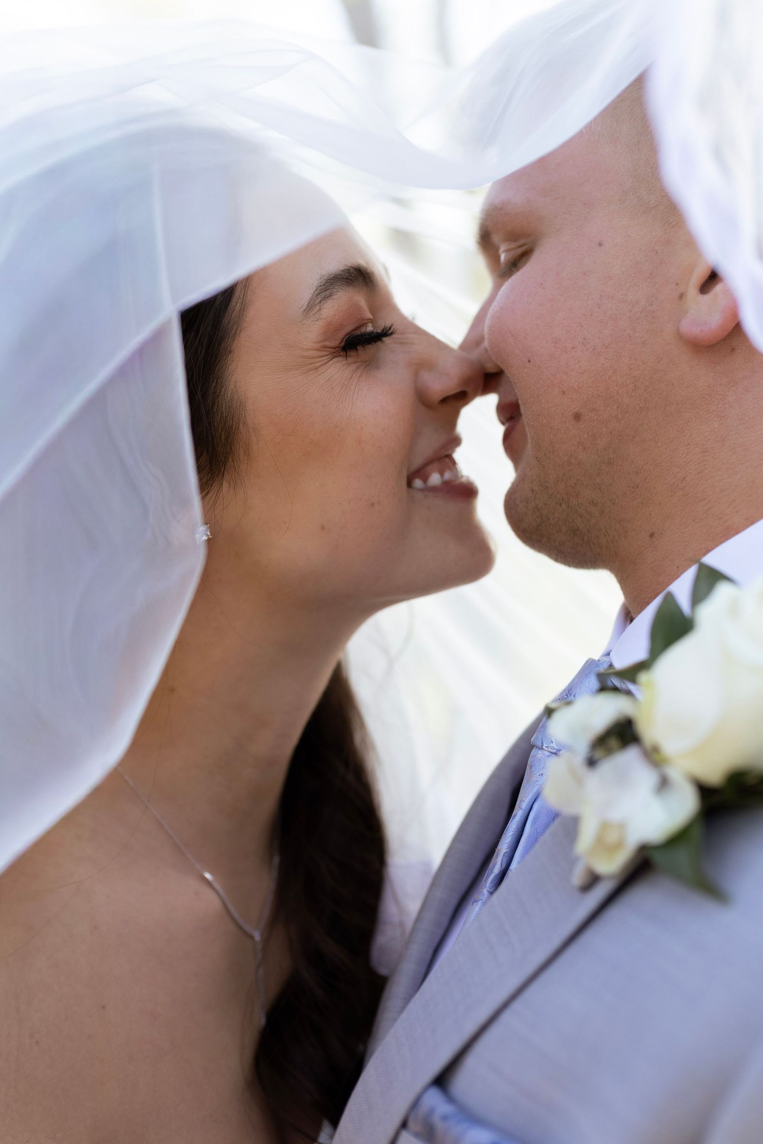 A bride and groom share a tender moment under a white veil, nearly kissing at their wedding ceremony.