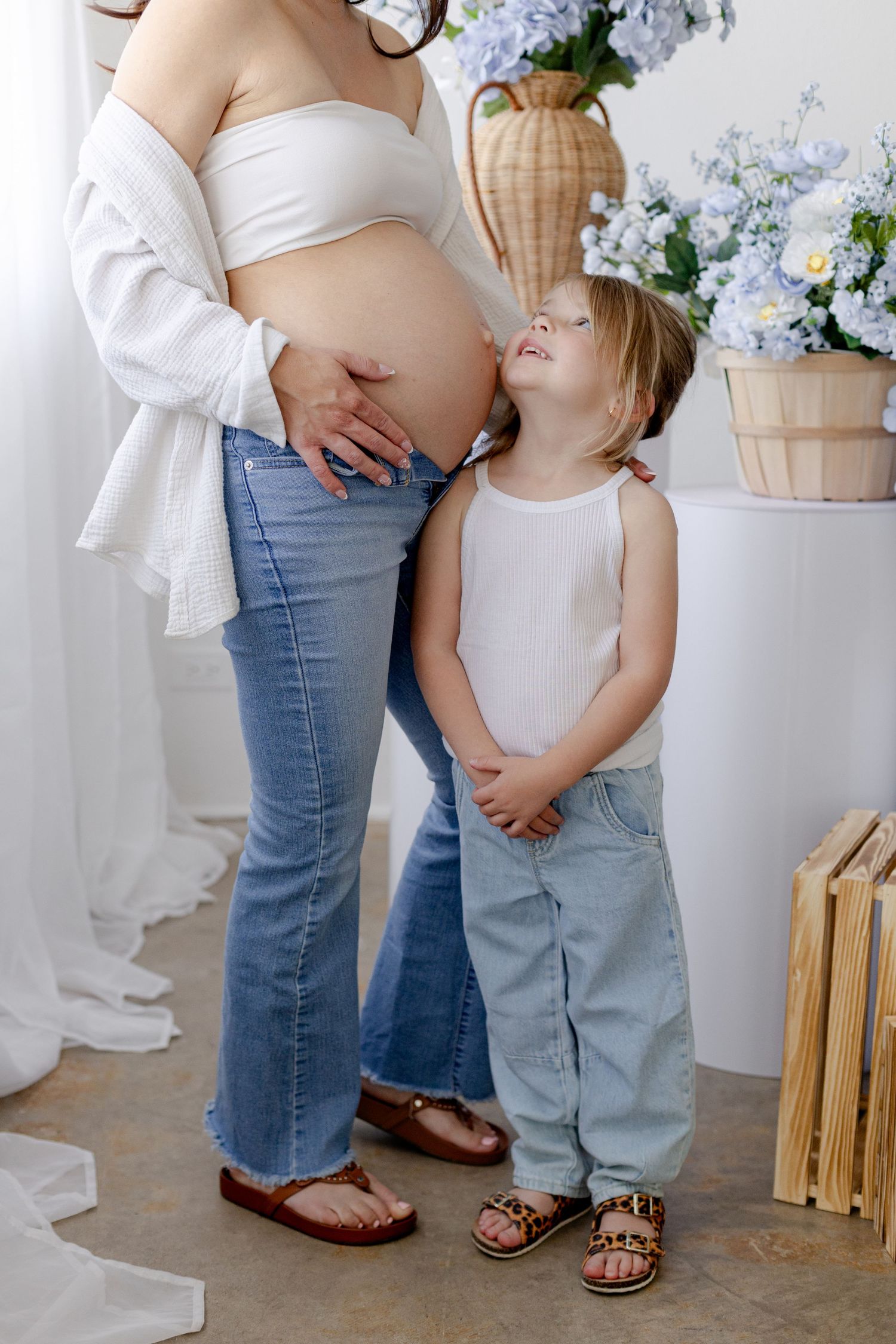 Young girl kisses pregnant mother's belly while looking up at her, surrounded by blue hydrangea flowers in bright studio.