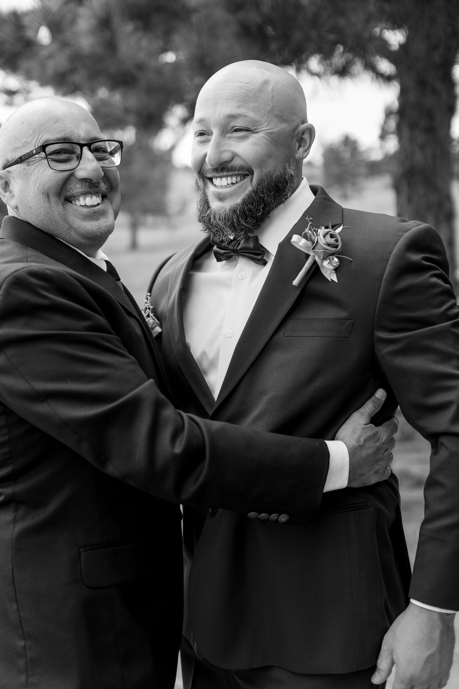 Two men in formal suits smiling and embracing outdoors at a wedding celebration in black and white.