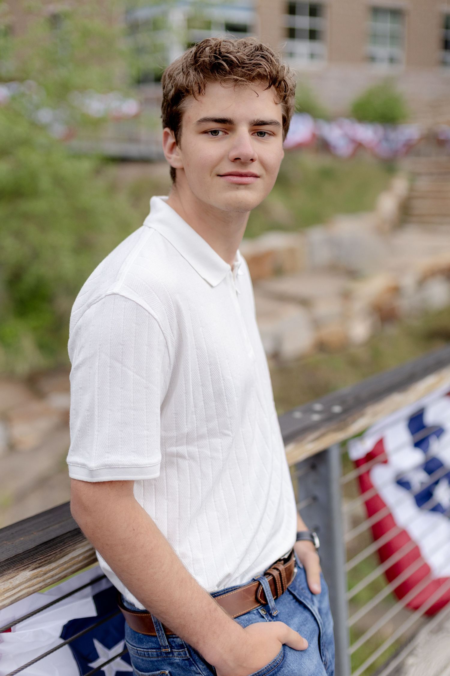 Young man in white polo shirt leaning on railing with American flags, smiling outdoors.