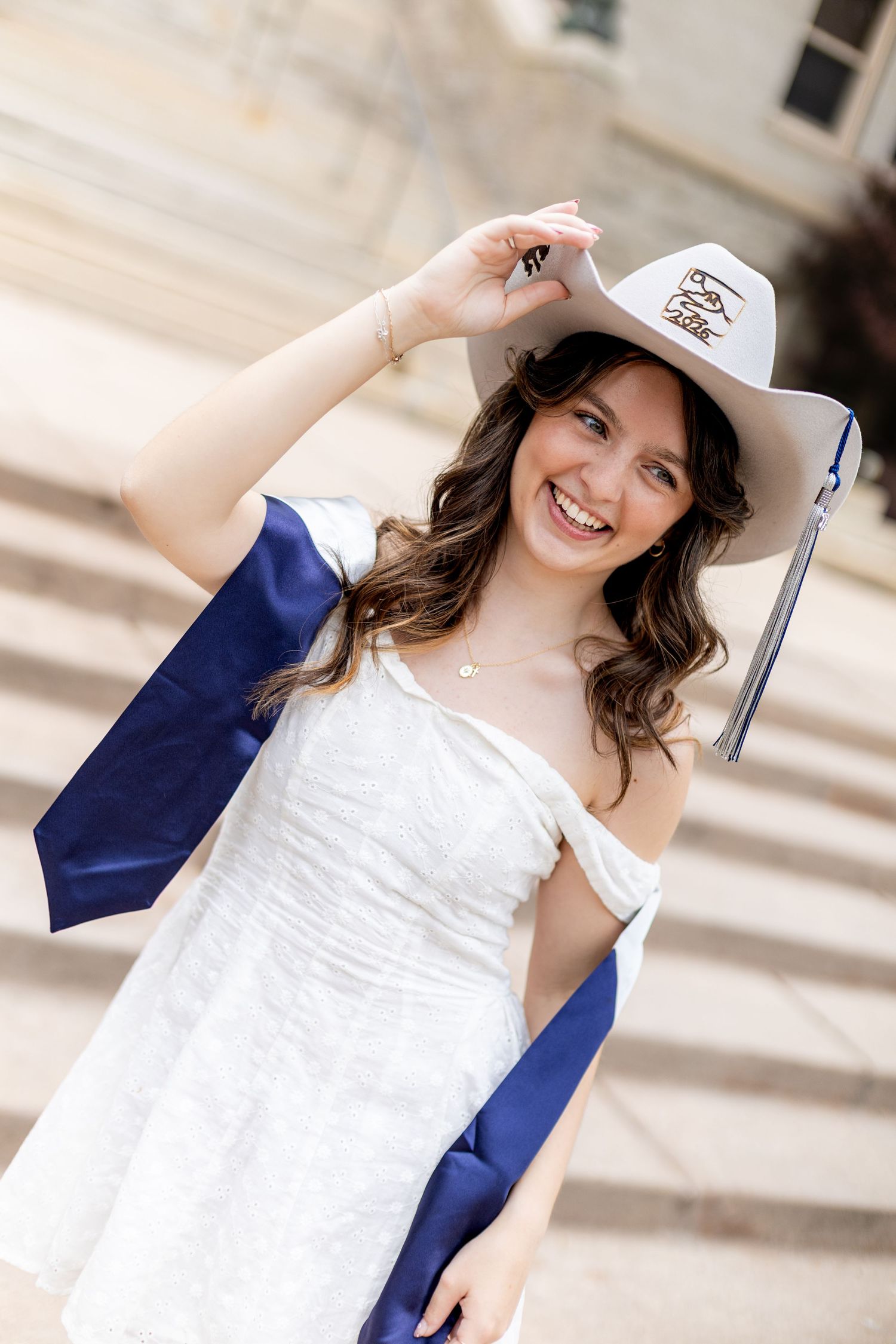 Smiling young woman in white dress and cowboy hat with blue graduation sash poses on steps for senior photos.