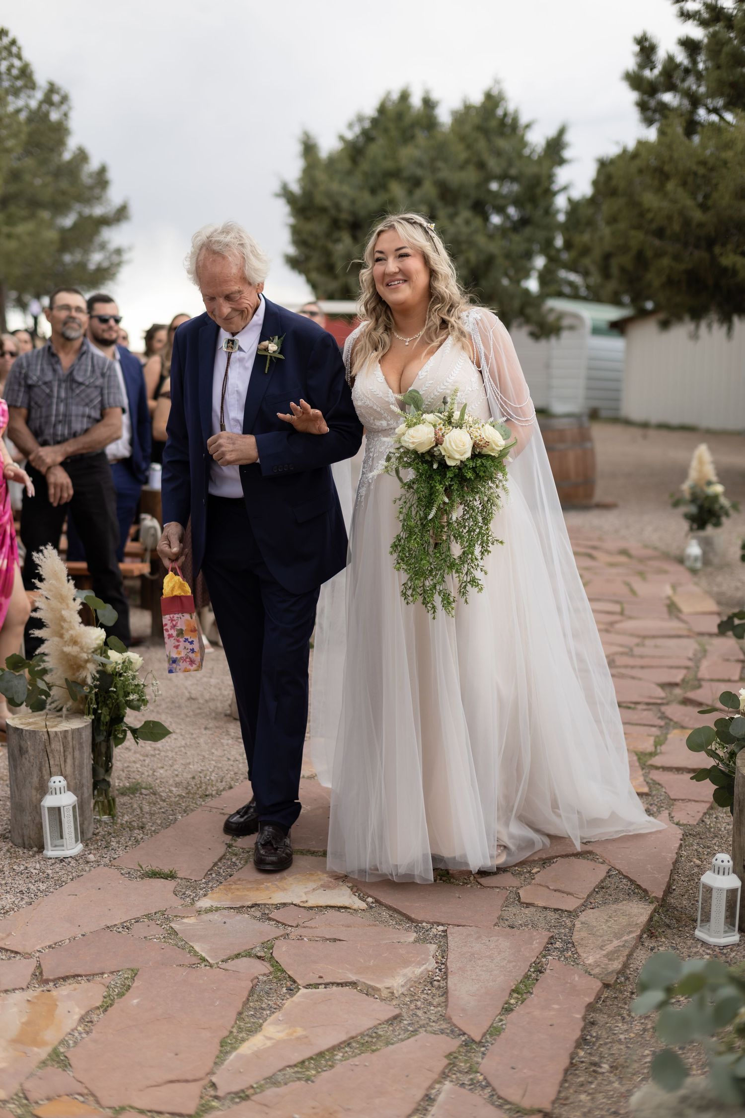 Smiling bride in white tulle gown holding bouquet walks down outdoor aisle with older man in navy suit.