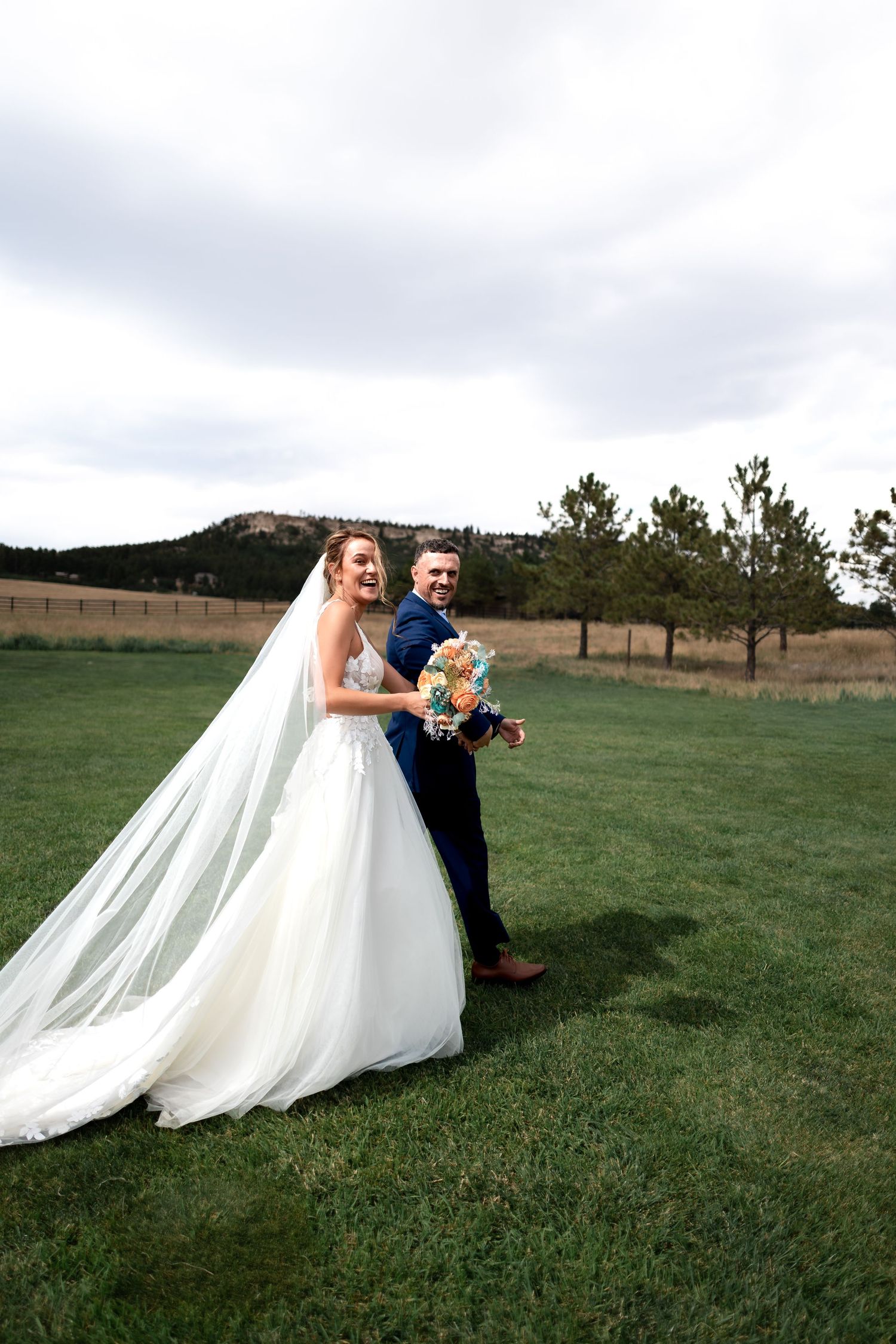 Bride and groom smiling on green lawn with mountain backdrop, bride's veil flowing in wind on wedding day.