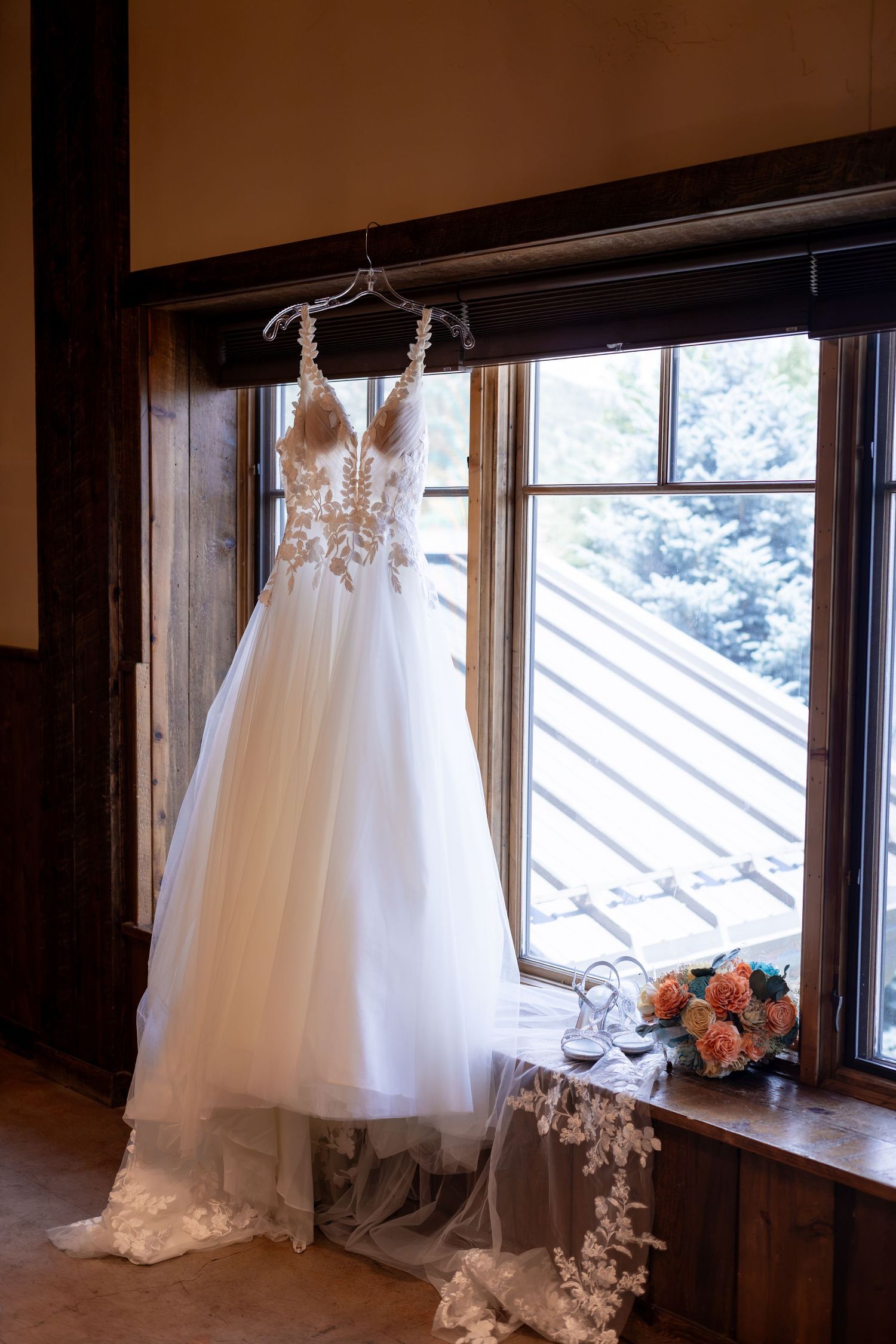 White lace wedding gown hanging by a window with a floral bouquet and bridal accessories on the sill.