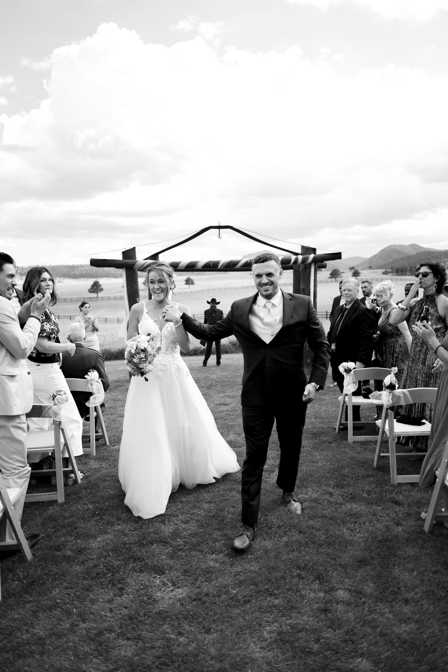Bride and groom walk down outdoor aisle after wedding ceremony in black and white photo.