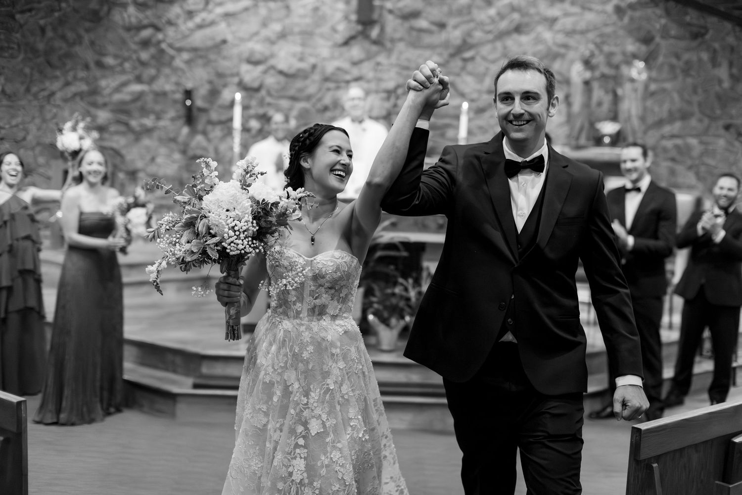 Joyful newlyweds celebrate recessional in black and white wedding photo, bride holding bouquet, groom in tuxedo.