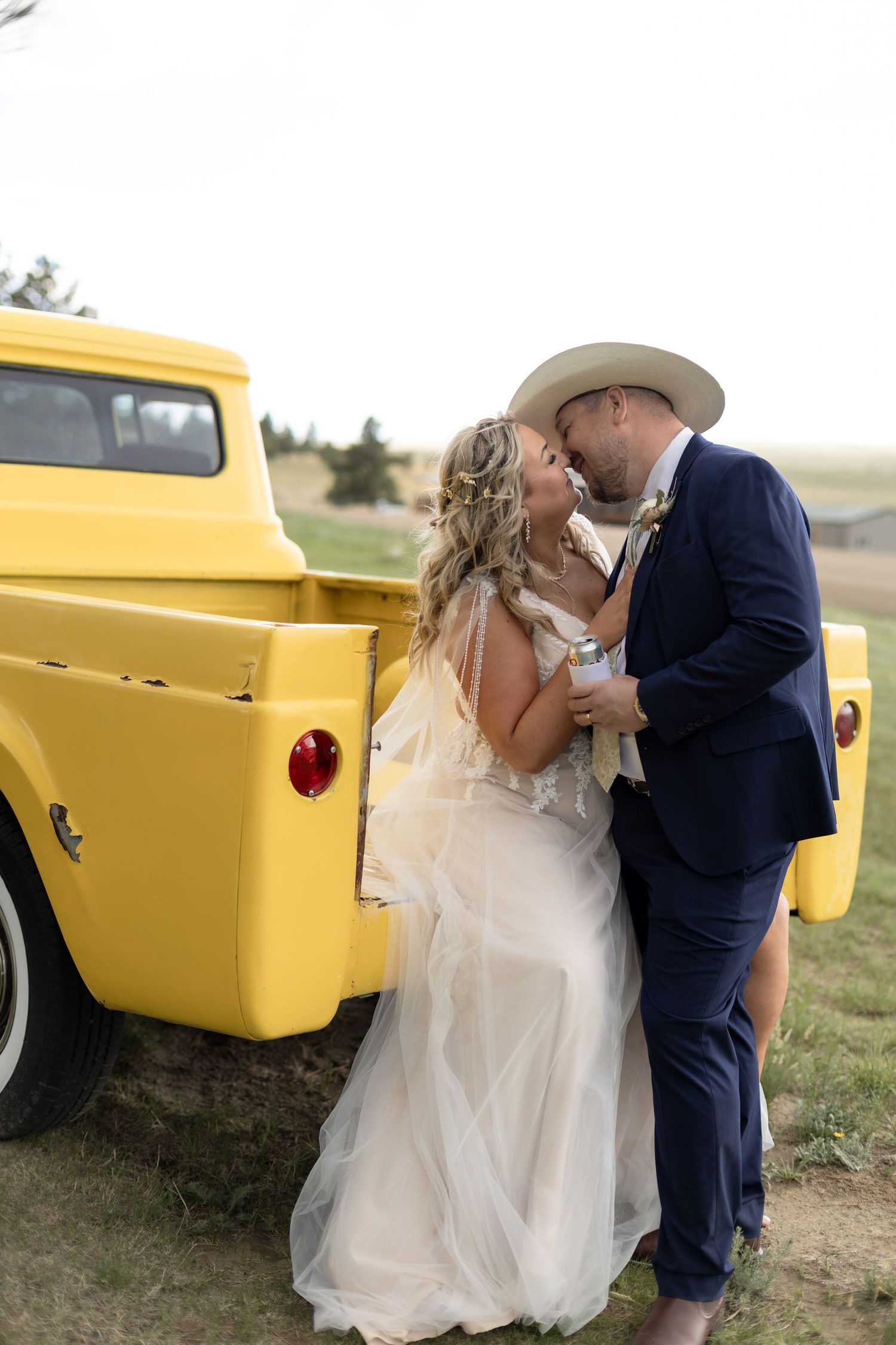 A bride and groom share a romantic kiss beside a vintage yellow pickup truck in a rustic outdoor wedding setting.