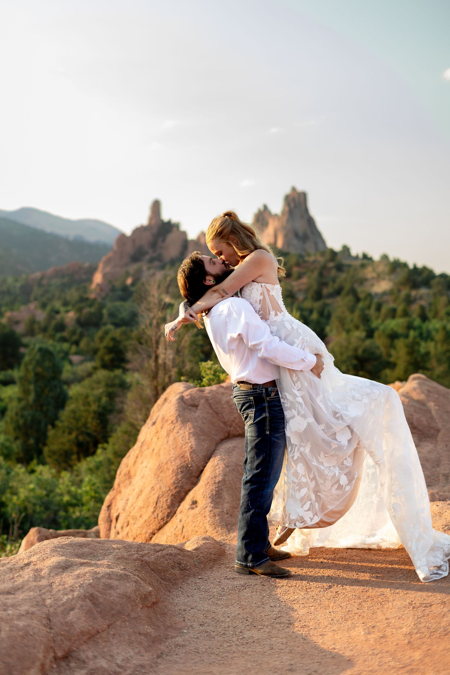 Couple sharing a romantic kiss at Garden of the Gods, Colorado, with red rock formations and green mountains in background.