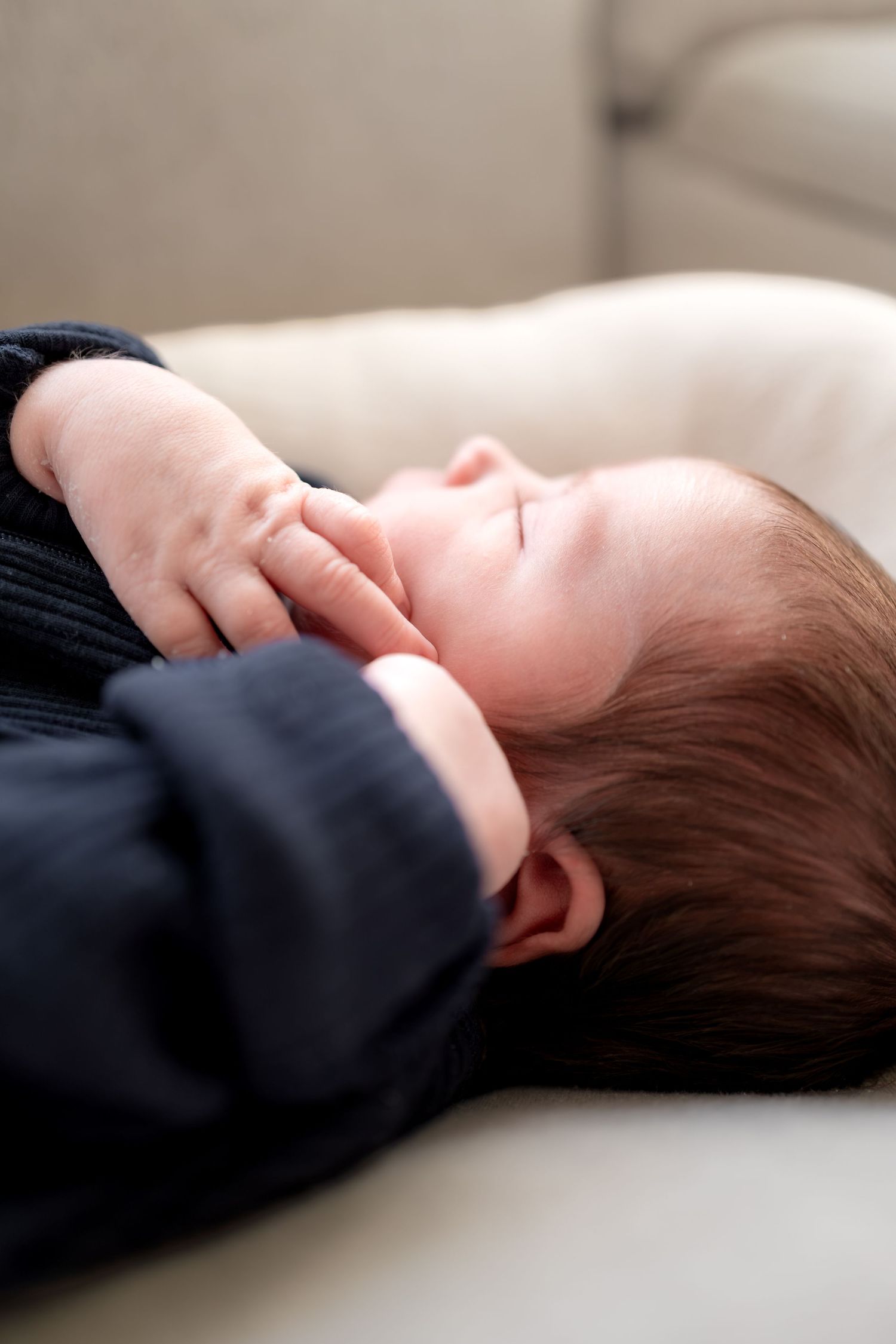 Peaceful newborn baby sleeping on a cream surface, wearing a navy outfit with tiny hand resting near face.