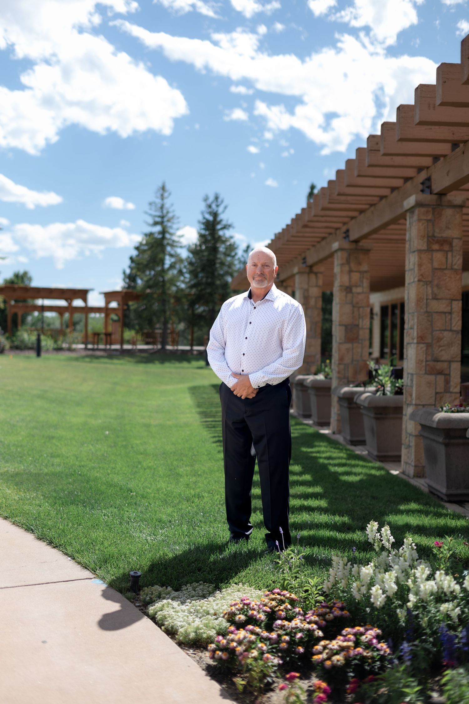 Man in white shirt and black pants standing in a beautifully landscaped garden with stone building and pergola.