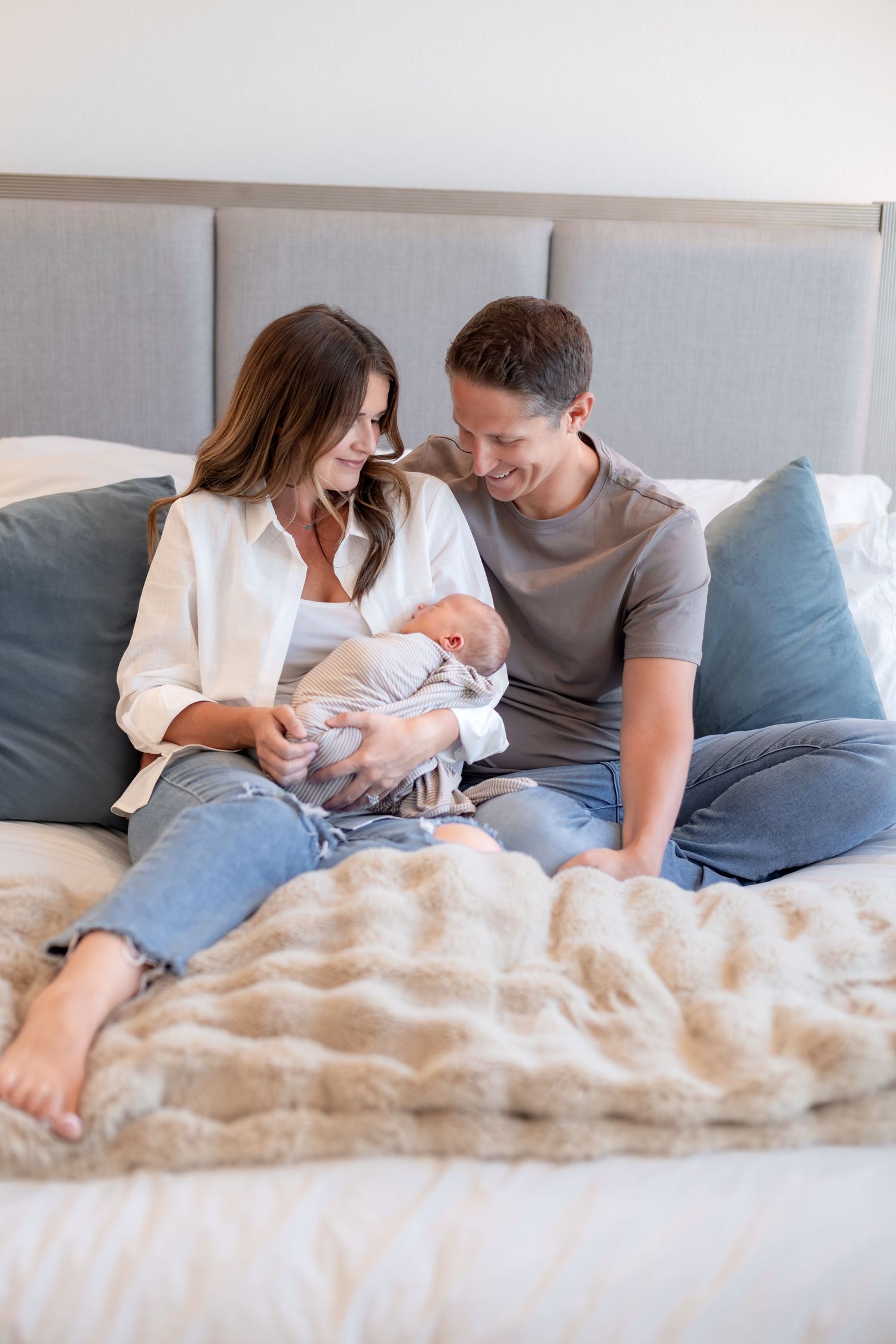 Happy couple sitting on a bed together, smiling as they hold their newborn baby.