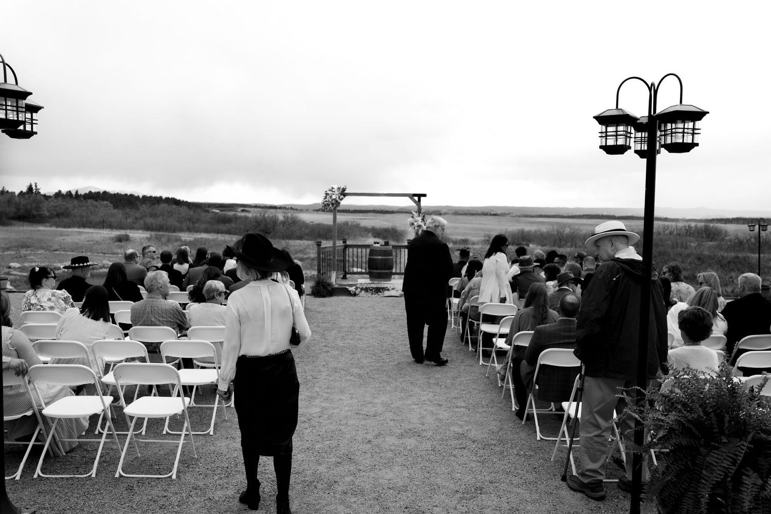 Black and white outdoor wedding ceremony with guests seated on folding chairs, officiant at altar, open field backdrop.
