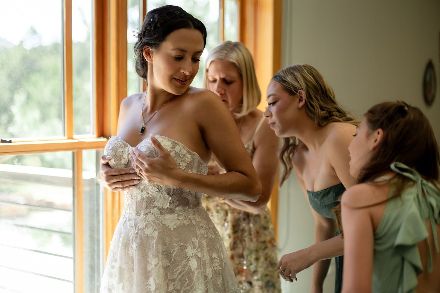 Bride in lace strapless wedding dress being helped by bridesmaids in sage green near a bright window.