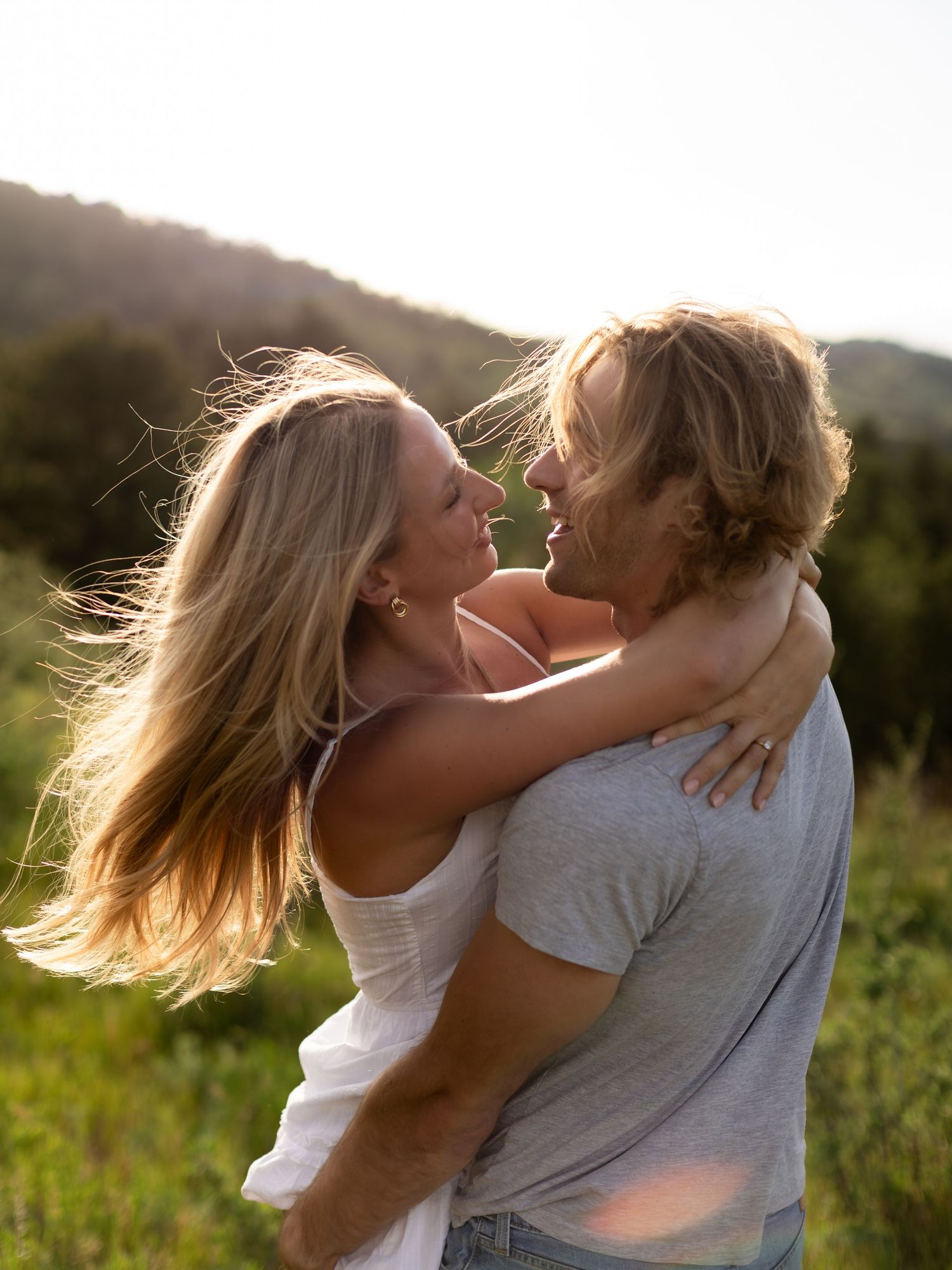 A couple embraces and gazes at each other in a sunlit meadow with hills in the background.